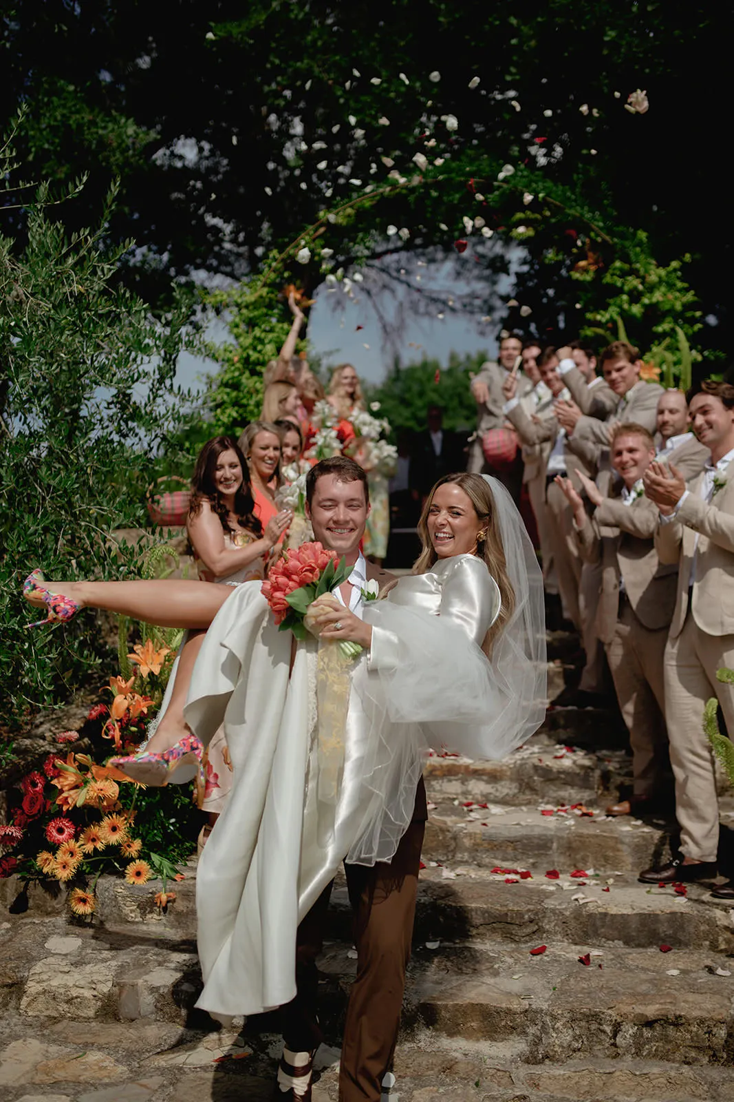 A joyful bride and groom celebrate outdoors in Tuscany as the groom carries the bride in his arms. The bride smiles, holding a bouquet, while guests and the wedding photographer capture every moment beneath a sunlit green archway. Tuscan Wedding Photographer