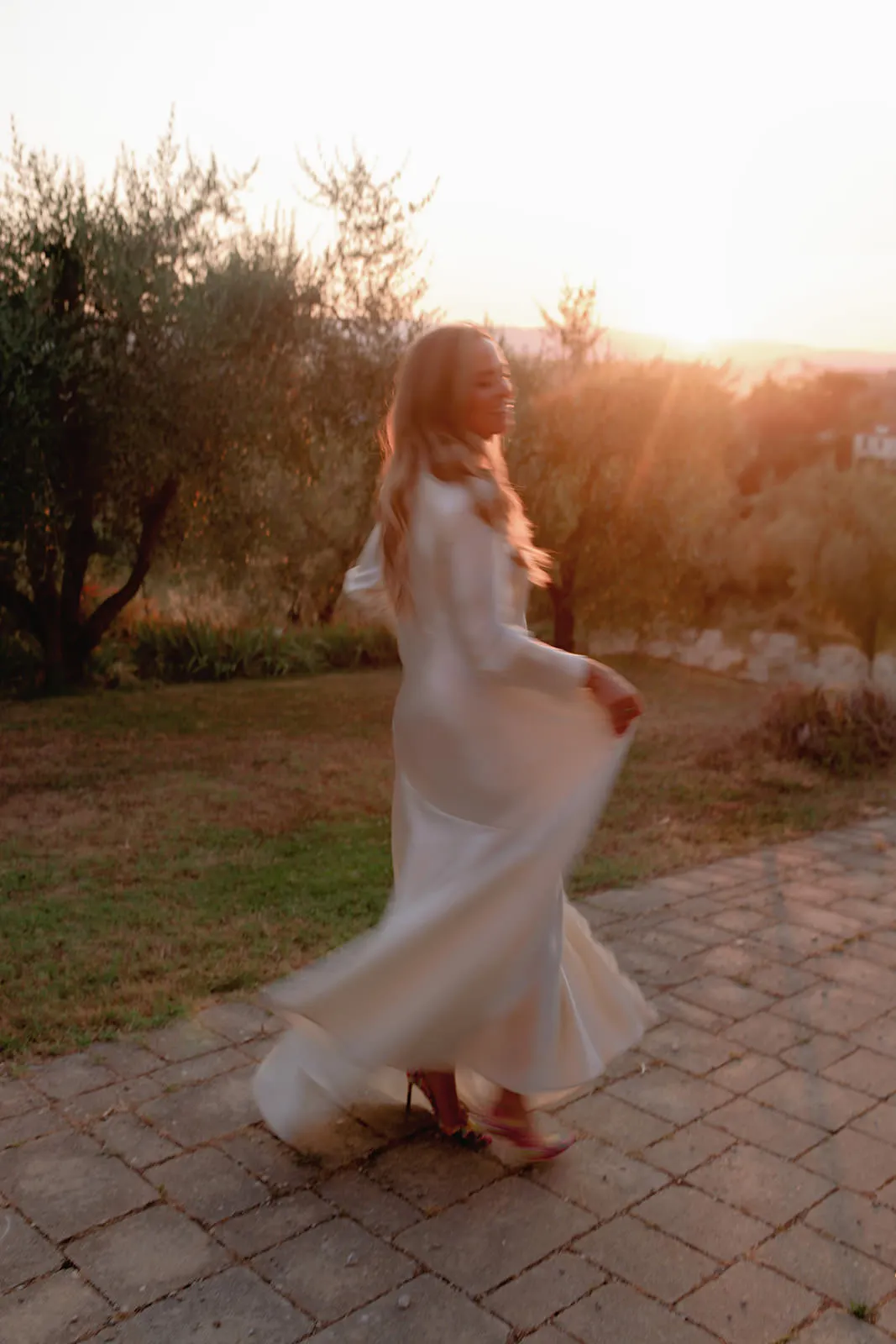 A woman in a long, flowing white dress twirls joyfully on a stone patio at sunset, as a wedding photographer in Tuscany captures her blurred motion. Sunlight streams through trees, casting a warm golden glow over the enchanting outdoor scene. Tuscan Wedding Photographer