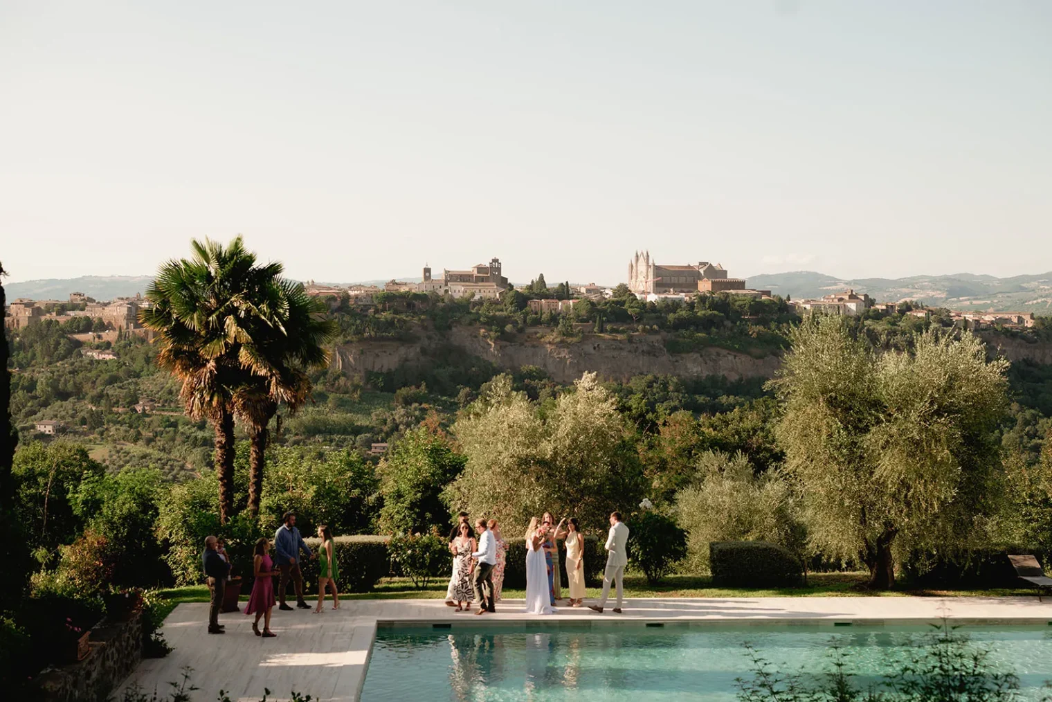 A small group in formal attire gathers beside a pool surrounded by greenery. A scenic hilltop town with historic buildings appears in the background—an idyllic setting for a wedding photographer Tuscany to capture timeless moments among palm and olive trees. Tuscan Wedding Photographer