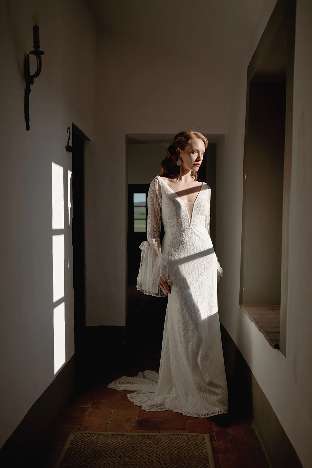 A woman in a white lace wedding dress with long sheer sleeves stands in a sunlit hallway, looking out a window. Captured by a wedding photographer Tuscany, the calm and contemplative moment is enhanced by her wavy hair and bold red lipstick. Tuscan Wedding Photographer