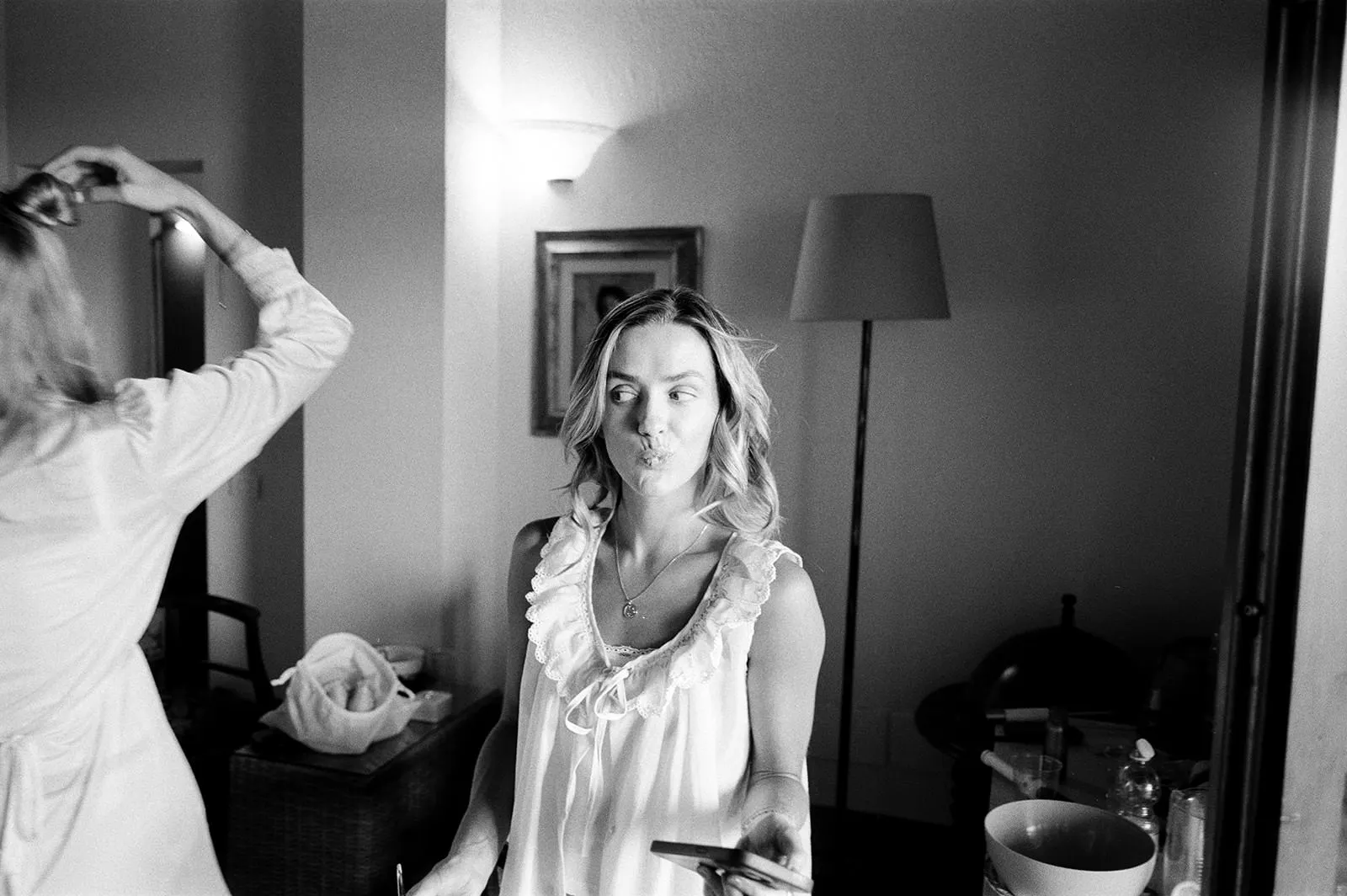 A woman in a ruffled sleeveless top stands indoors, making a playful puckered face at a mirror—like she's posing for a wedding photographer. She holds her phone, while another person adjusts their hair in the softly lit room filled with art and scattered items. Tuscan Wedding Photographer