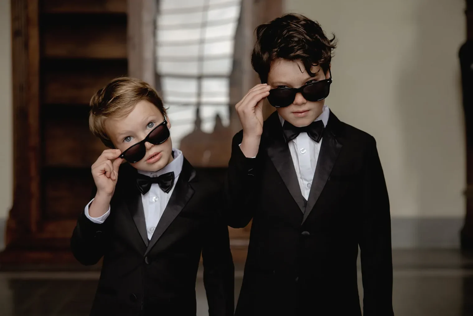 Two young boys in black tuxedos and bow ties stand indoors, each lifting sunglasses to look at the camera. Both have wavy hair; the taller one is on the right. Captured in a blurred interior with a wooden staircase by a wedding photographer Tuscany. Tuscan Wedding Photographer