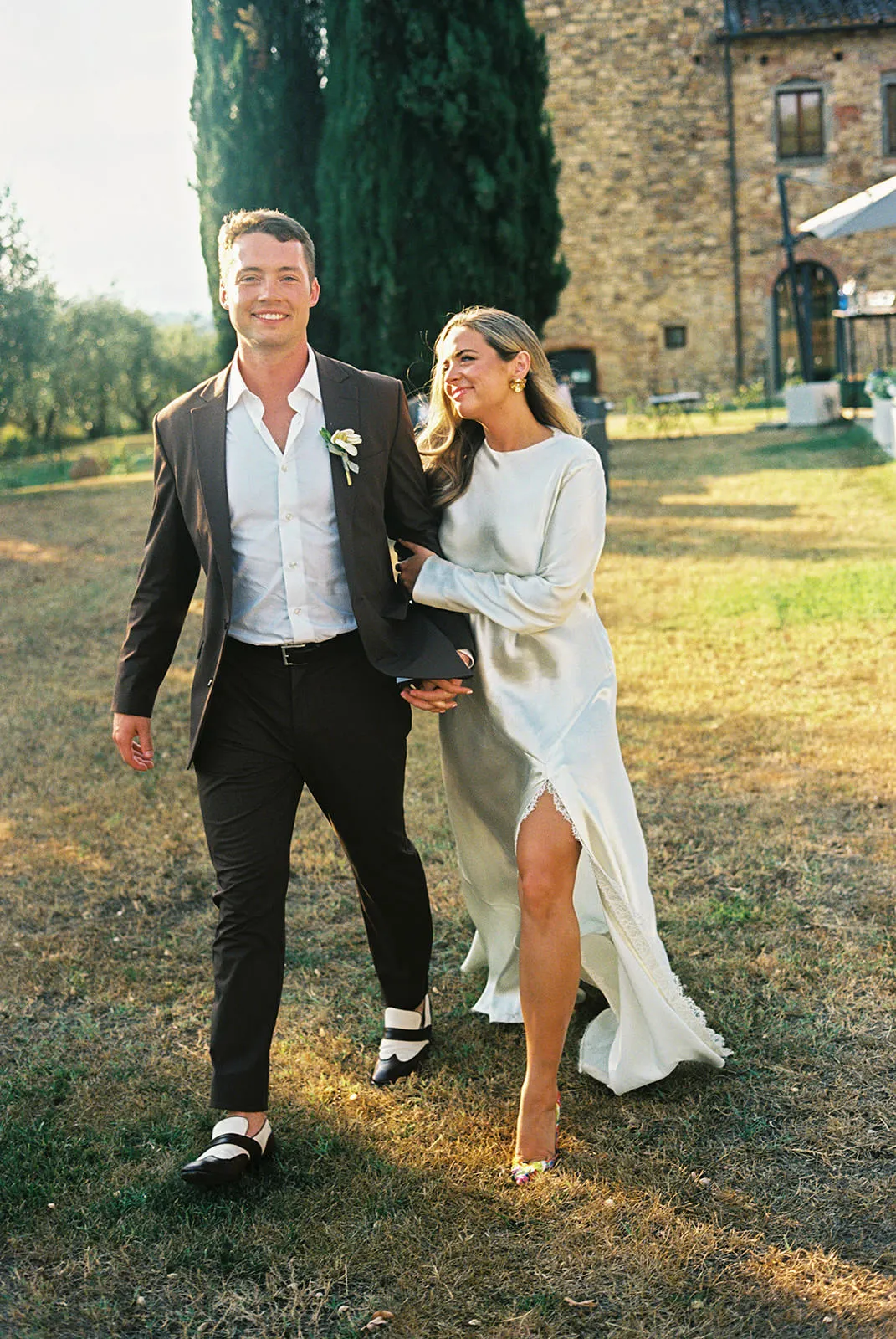 A smiling man in a black suit and white shirt walks holding hands with a woman in a long white dress on grass, outdoors, near trees and a stone building. She looks at him happily. Sunlight brightens the scene. Tuscan Wedding Photographer
