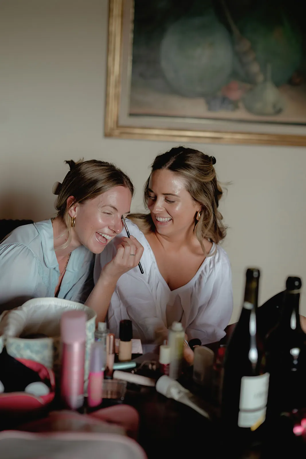 Two women sit together smiling as one applies makeup to the other’s face. They are surrounded by makeup products and bottles on a table. Both appear happy and relaxed, with a painting hanging on the wall behind them. The setting looks casual and friendly. Tuscan Wedding Photographer