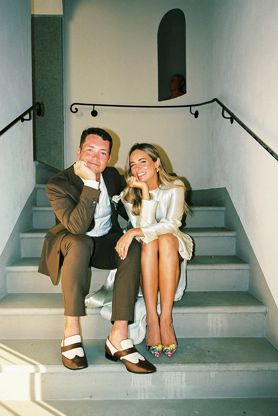 A smiling man in a brown suit and white heels sits on stairs with a woman in a silky light dress and colorful heels. Both rest their chins on their hands. Sunlight highlights them; a shadowy figure is in the background at the top of the stairs. Tuscan Wedding Photographer