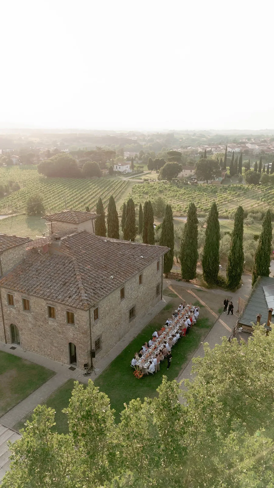 A group of people sits at a long outdoor table beside a rustic stone building, surrounded by trees and vineyards under a hazy sky, suggesting a celebration or meal in a scenic countryside setting. Tuscan Wedding Photographer