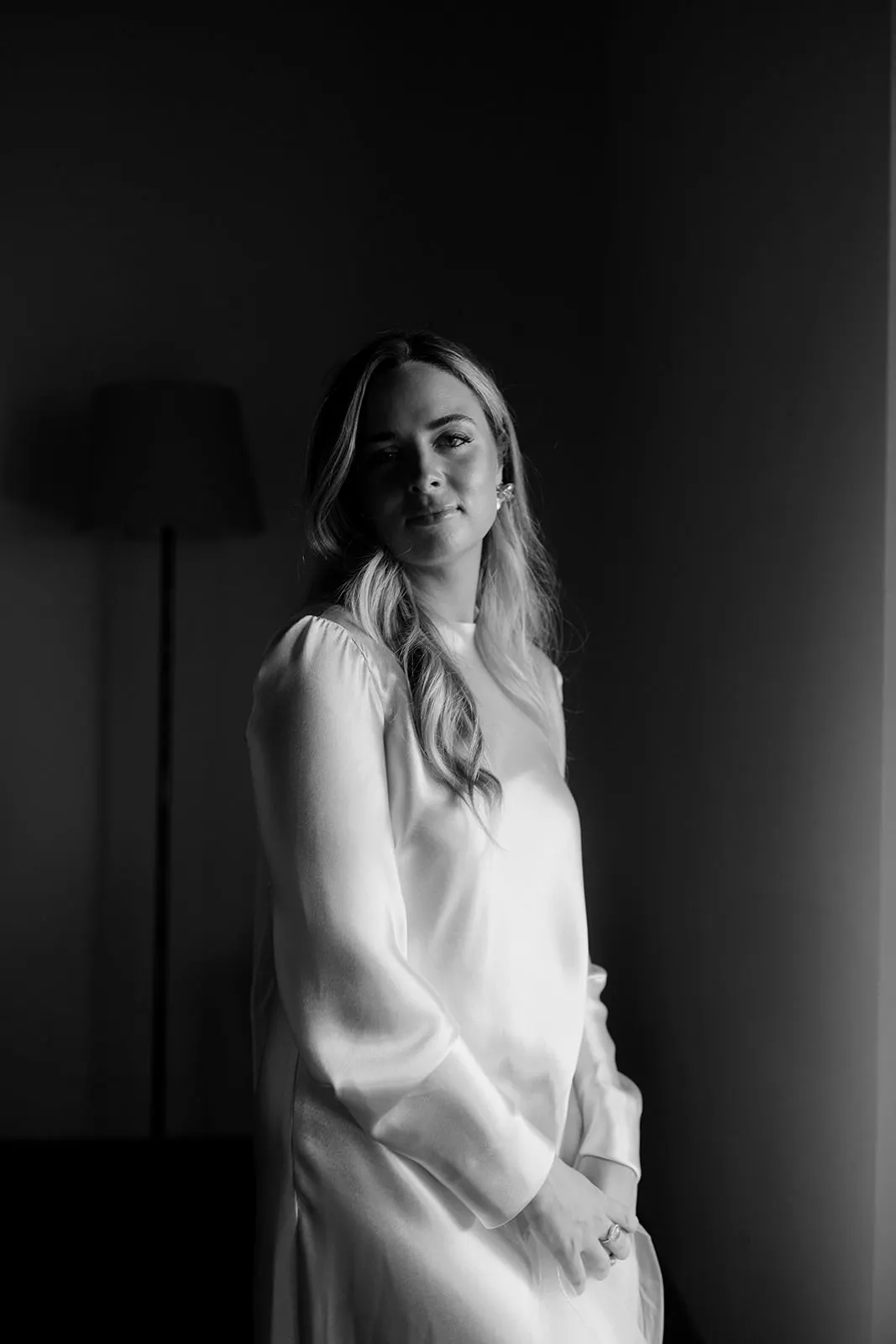A woman in a long-sleeved, white dress stands by a window, bathed in soft natural light. Her hair is loose and wavy. The room is dimly lit, and a floor lamp appears in the background. The photo is black and white. Tuscan Wedding Photographer