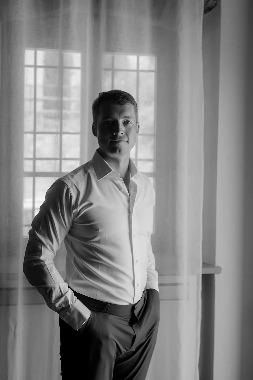 A man stands indoors by a window with sheer curtains, wearing a white button-up shirt and dark pants. He has one hand in his pocket and is looking at the camera, softly lit by natural light coming through the window. The photo is black and white. Tuscan Wedding Photographer