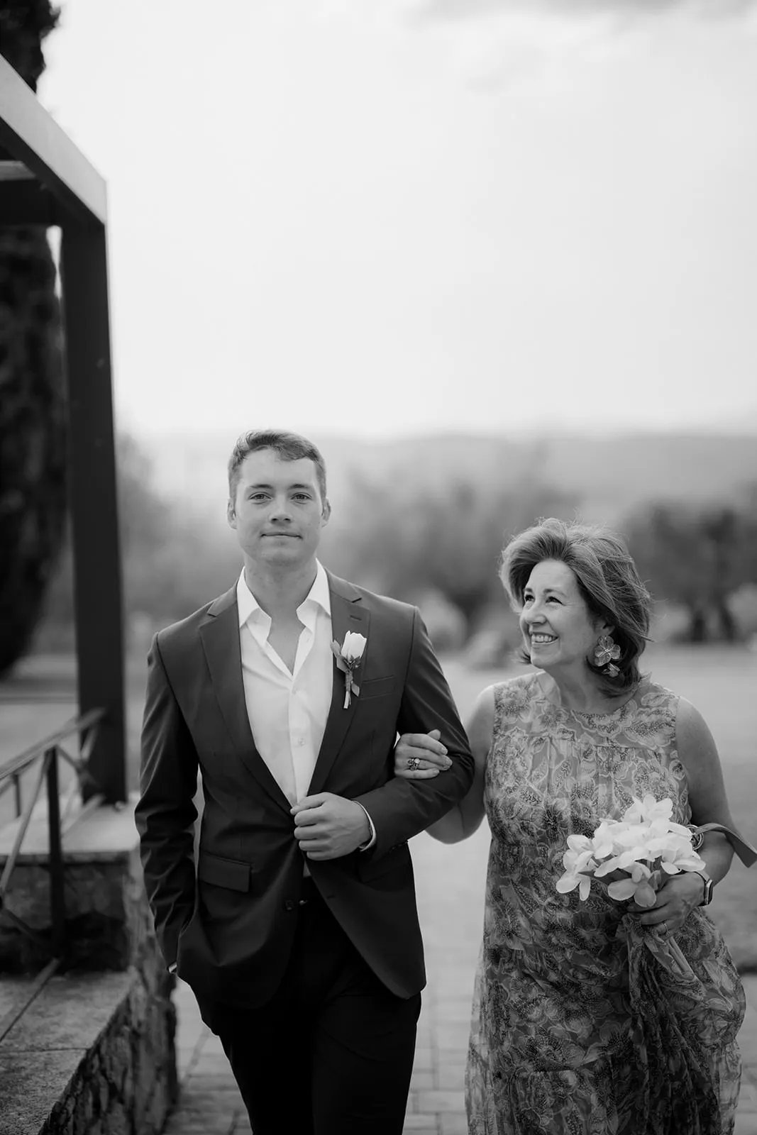 A young man in a suit walks arm-in-arm with an older woman in a floral dress holding a bouquet. They are outdoors on a stone path, with blurred trees and hills in the background. The woman is smiling at him fondly, while he looks ahead. The image is black and white. Tuscan Wedding Photographer