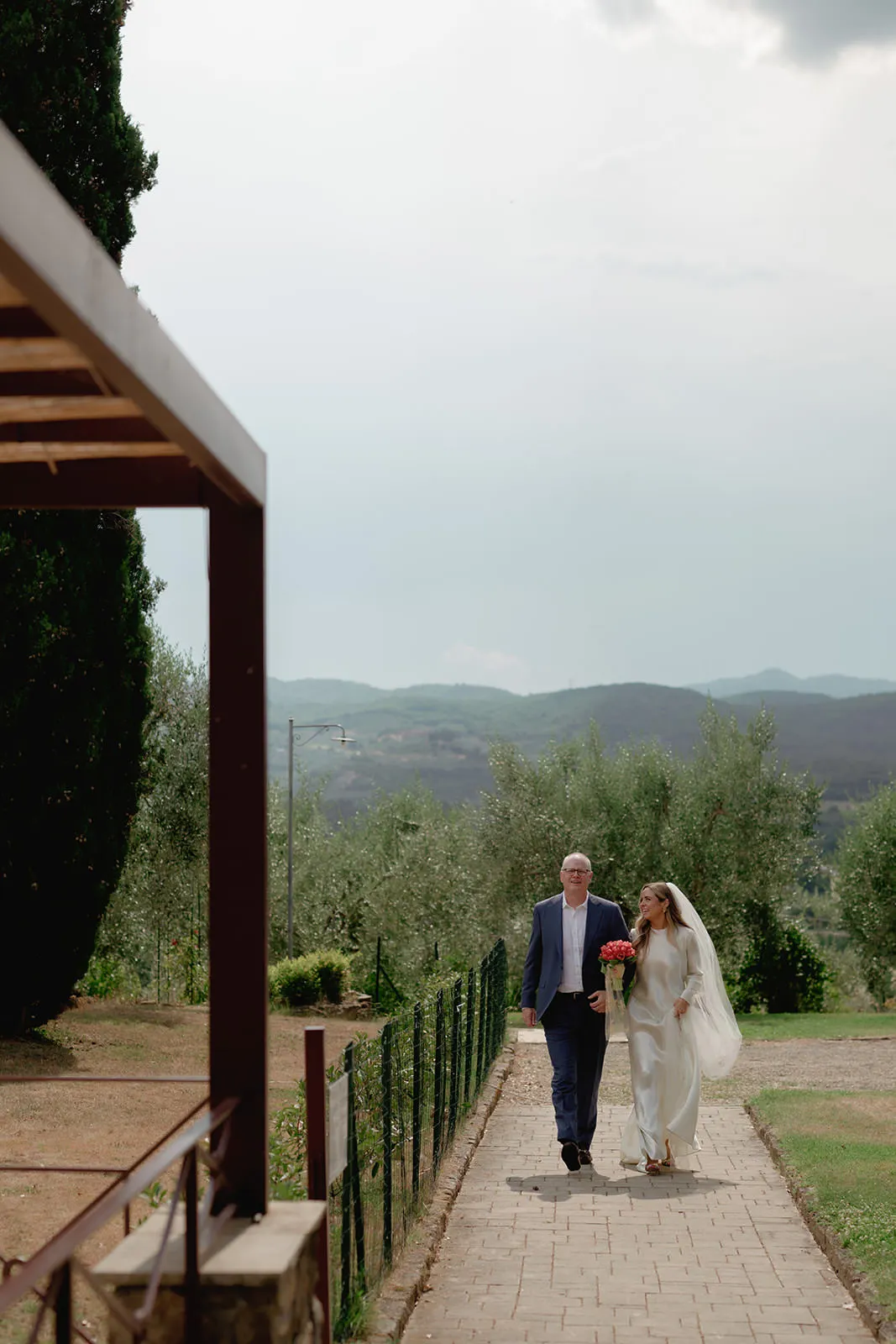A bride in a white dress and veil walks beside an older man in a blue suit along a garden path, holding a bouquet. Green hills and trees are in the background under a cloudy sky. Part of a wooden structure is visible on the left. Tuscan Wedding Photographer