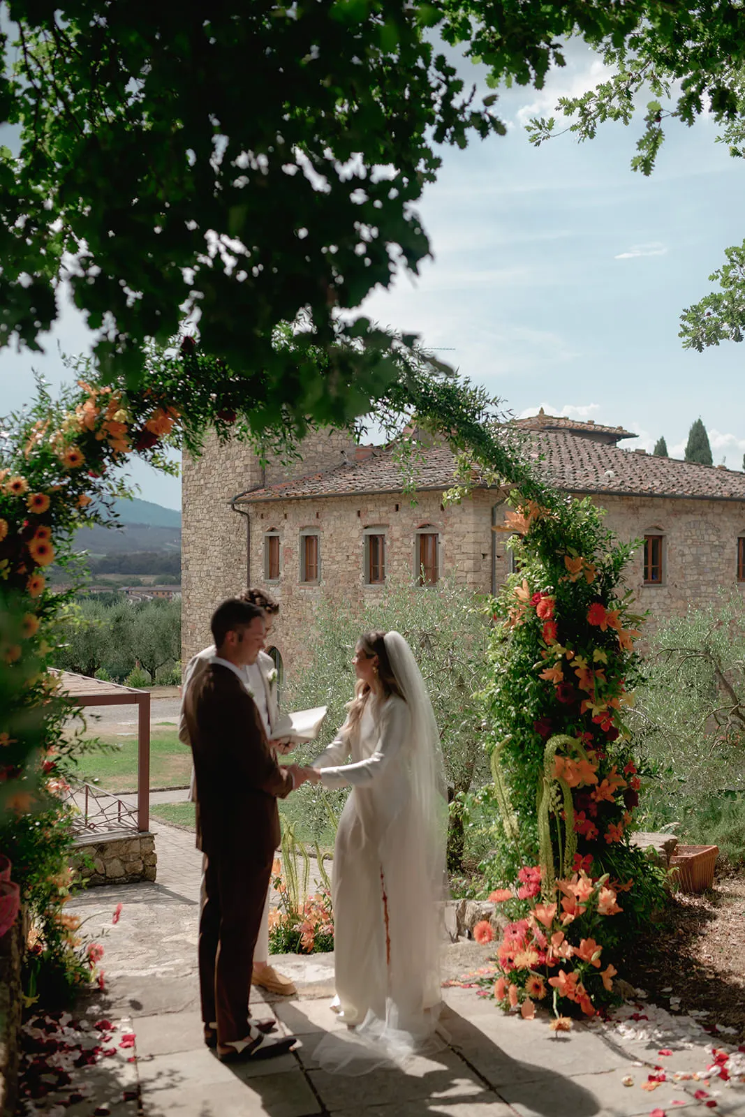 A bride and groom stand under a flower-adorned arch, exchanging vows outdoors. The bride wears a white dress and veil, while the groom wears a brown suit. Behind them is a stone building, greenery, and distant hills under a partly cloudy sky. Tuscan Wedding Photographer