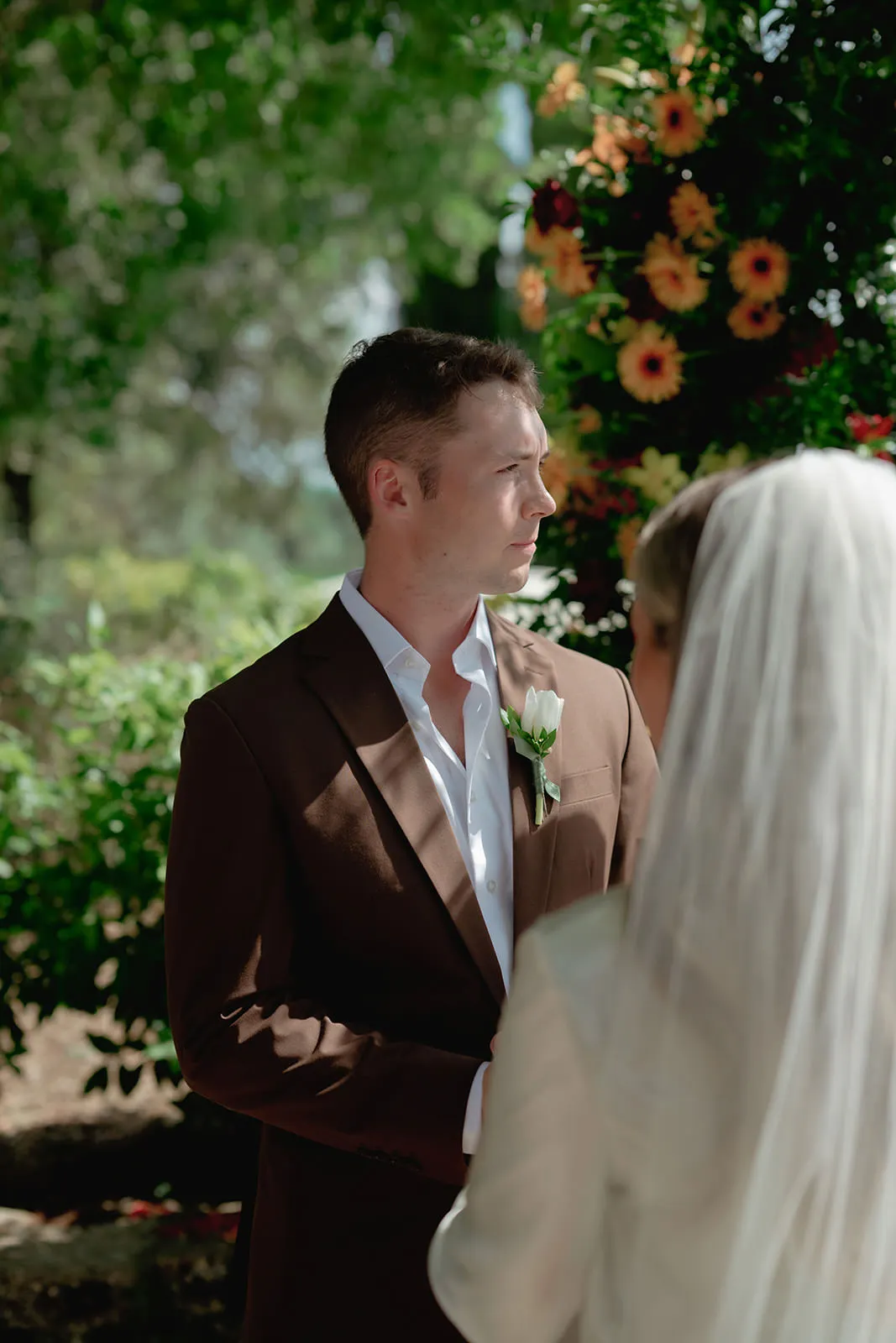 A groom in a brown suit stands facing a bride in a white veil. They are outdoors, surrounded by greenery and yellow flowers, with sunlight shining on them. Both appear focused and serious, likely during a wedding ceremony. Tuscan Wedding Photographer