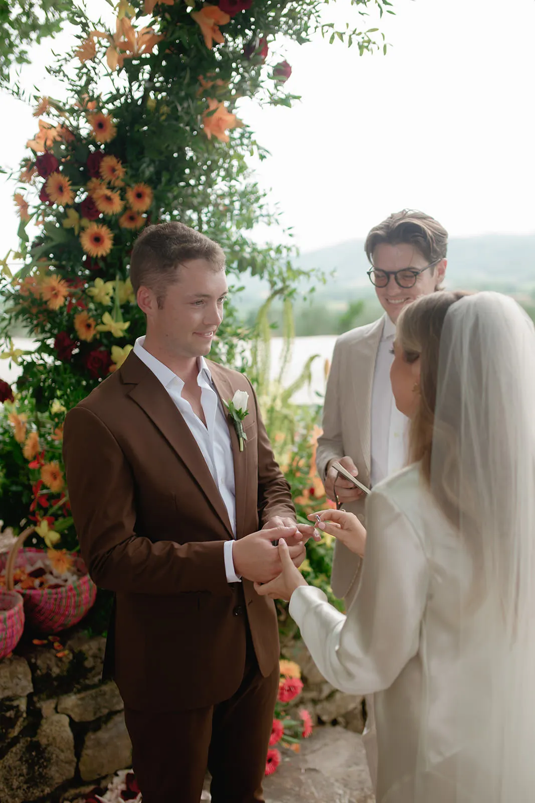 A groom in a brown suit and a bride in a veil exchange rings during an outdoor wedding ceremony. An officiant in a beige suit stands behind them. Colorful flowers decorate the archway, with green hills and trees visible in the background. Tuscan Wedding Photographer