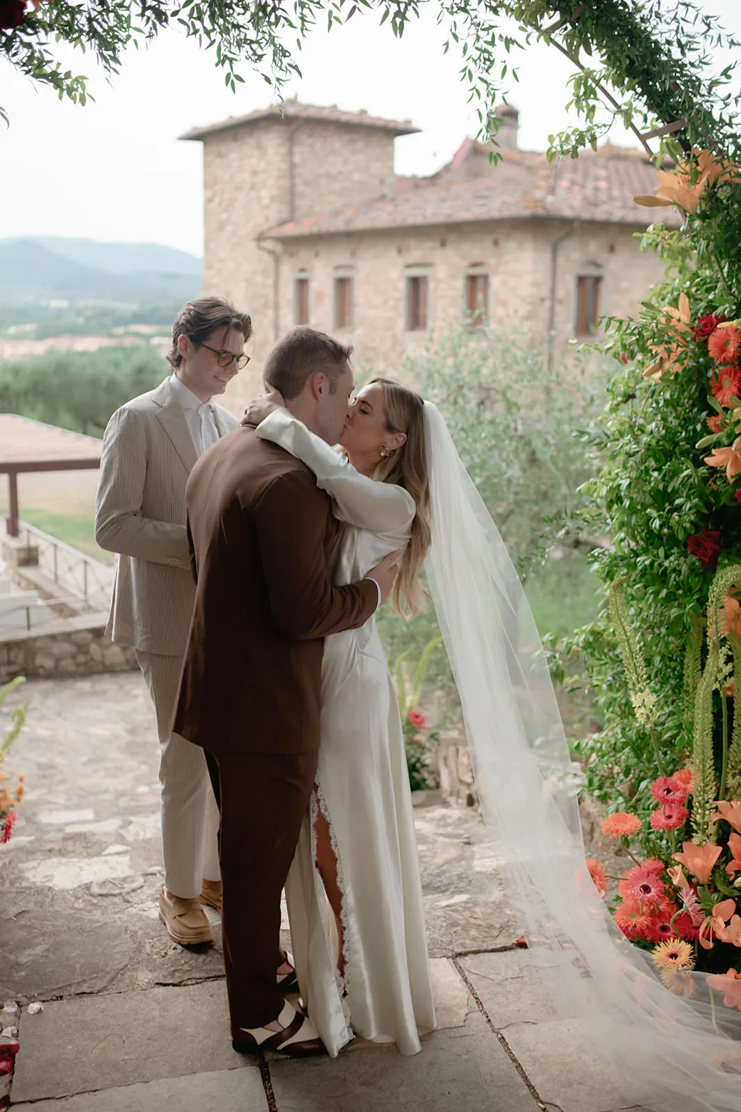 A bride in a white dress and veil kisses the groom in a brown suit during an outdoor wedding ceremony. A person in a tan suit stands behind them. Bright flowers and greenery frame the scene, with a stone building and hills in the background. Tuscan Wedding Photographer