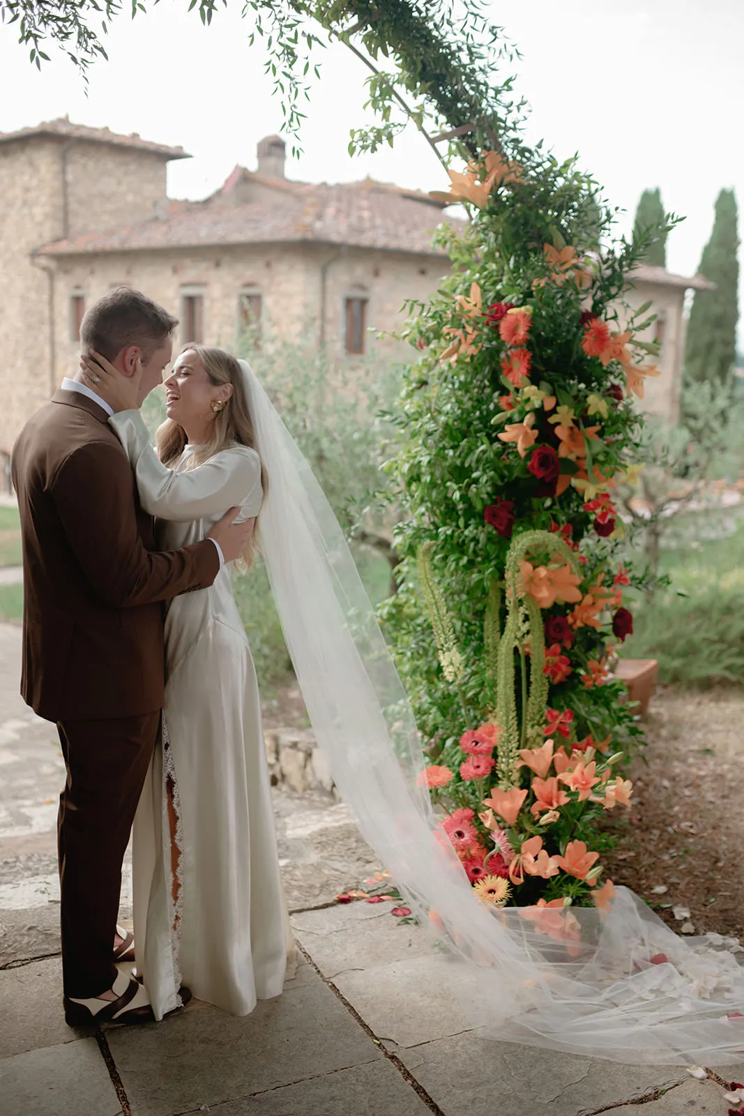 A bride in a long white dress and veil embraces a groom in a brown suit under a floral archway, adorned with vibrant orange and coral flowers, outside a rustic stone building surrounded by greenery. Both look lovingly at each other, smiling softly. Tuscan Wedding Photographer