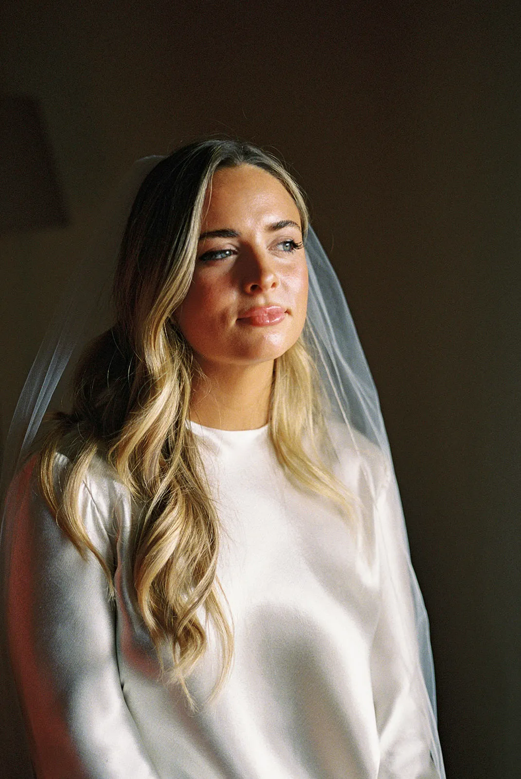 A woman with wavy blonde hair wears a white satin dress and a sheer veil, gazing softly to the side. The lighting highlights her face and hair, giving a warm, serene feeling. The background is dark and out of focus. Tuscan Wedding Photographer