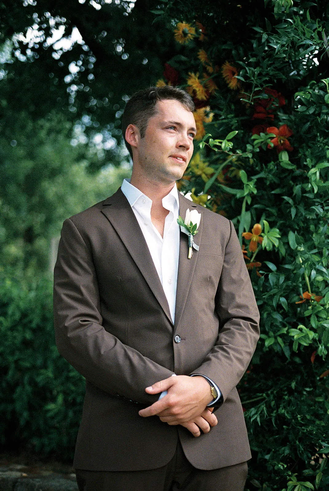 A man in a brown suit and white shirt stands outdoors, hands clasped, wearing a white boutonniere. He looks slightly to the side with a thoughtful expression. Green foliage and orange flowers fill the background, suggesting a garden or outdoor event. Tuscan Wedding Photographer