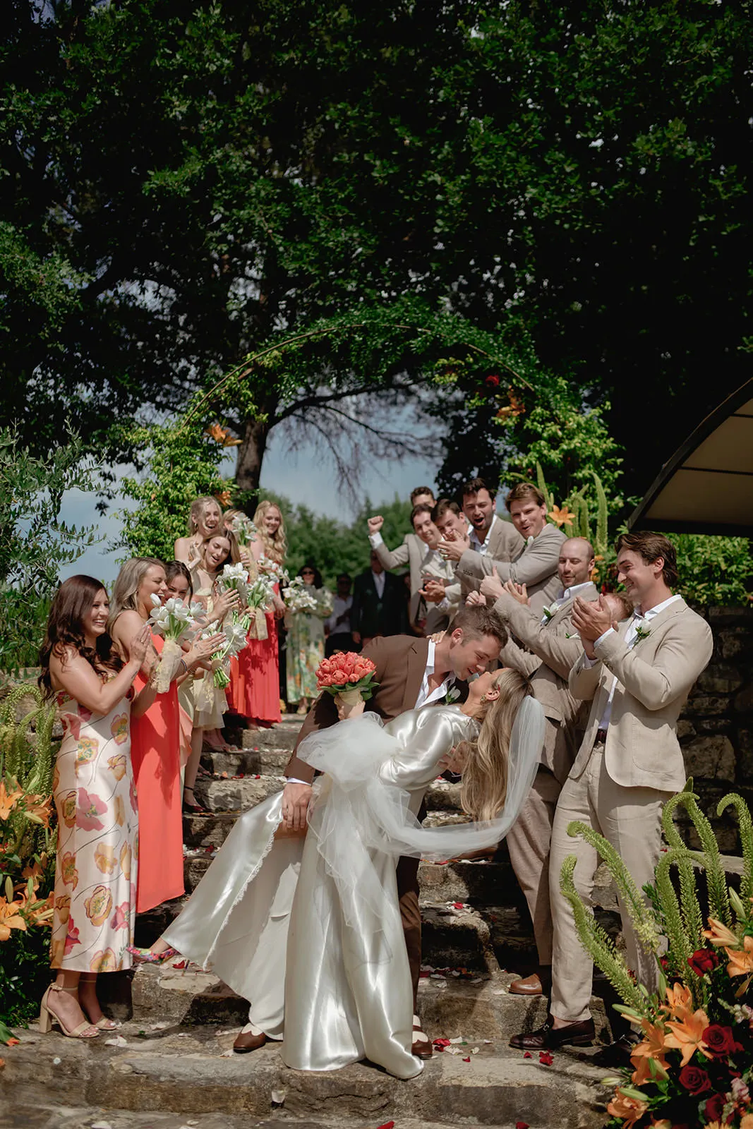 A groom dips and kisses the bride on stone steps, surrounded by cheering and clapping wedding guests in light suits and coral dresses, with vibrant flowers and greenery all around. The scene is bright, joyful, and outdoors under a lush green arch. Tuscan Wedding Photographer
