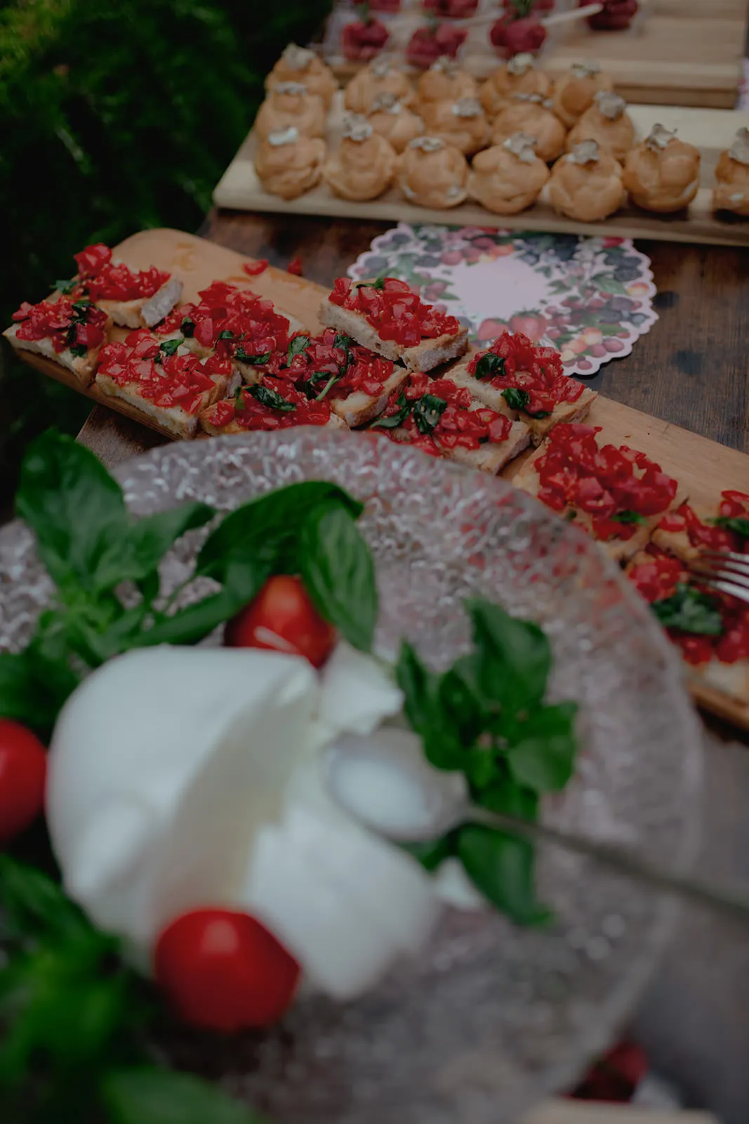 A close-up of a table with bruschetta topped with diced tomatoes and herbs, a glass bowl with fresh mozzarella, basil leaves, and cherry tomatoes, and cream puffs on a wooden board in the background. The scene is set on a rustic wooden surface. Tuscan Wedding Photographer