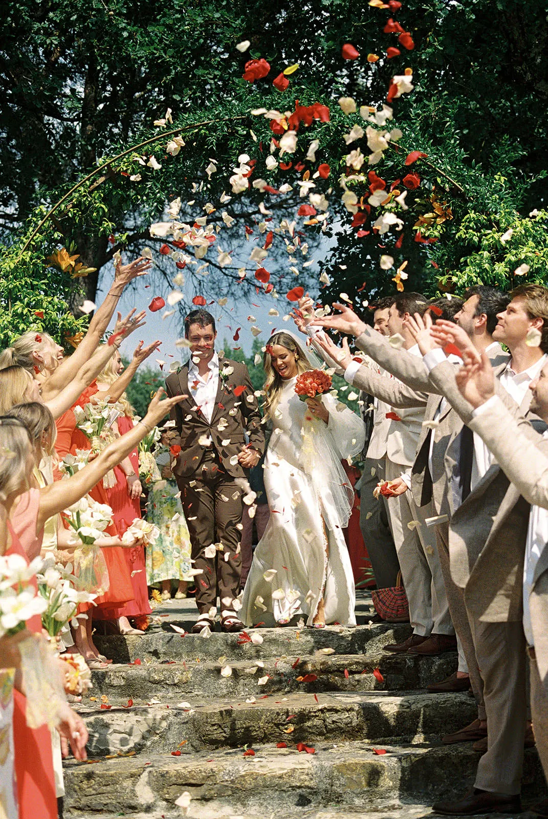 A bride in a white dress and groom in a brown suit walk down stone steps, smiling, as guests in peach and beige outfits throw flower petals in the air under a leafy green arch on a sunny day. Tuscan Wedding Photographer