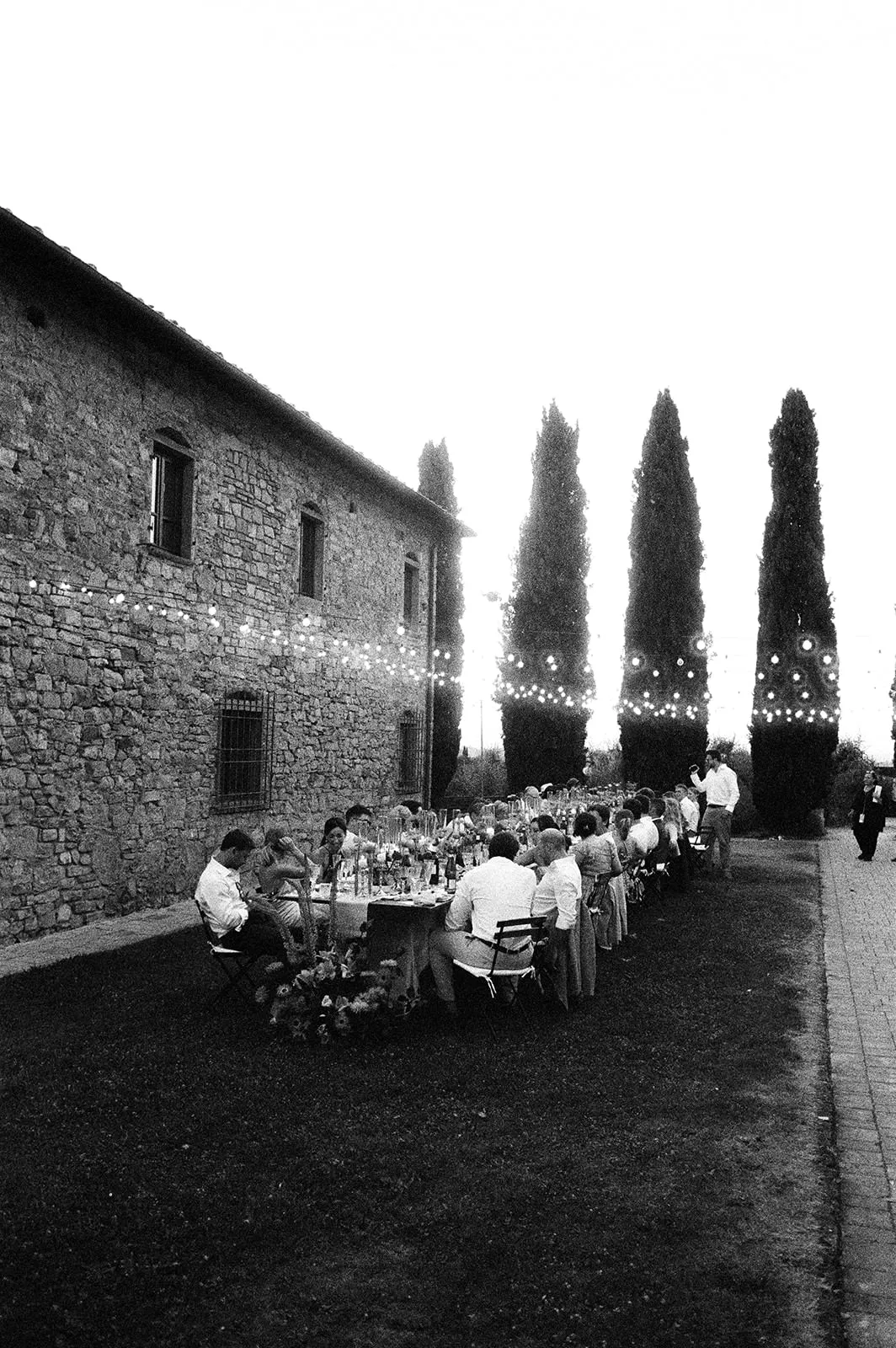 A group of people sit at a long outdoor dining table beside a stone building. String lights hang above. Tall cypress trees line the background. The scene is festive and elegant, with guests dressed formally and engaged in conversation. The photo is black and white. Tuscan Wedding Photographer