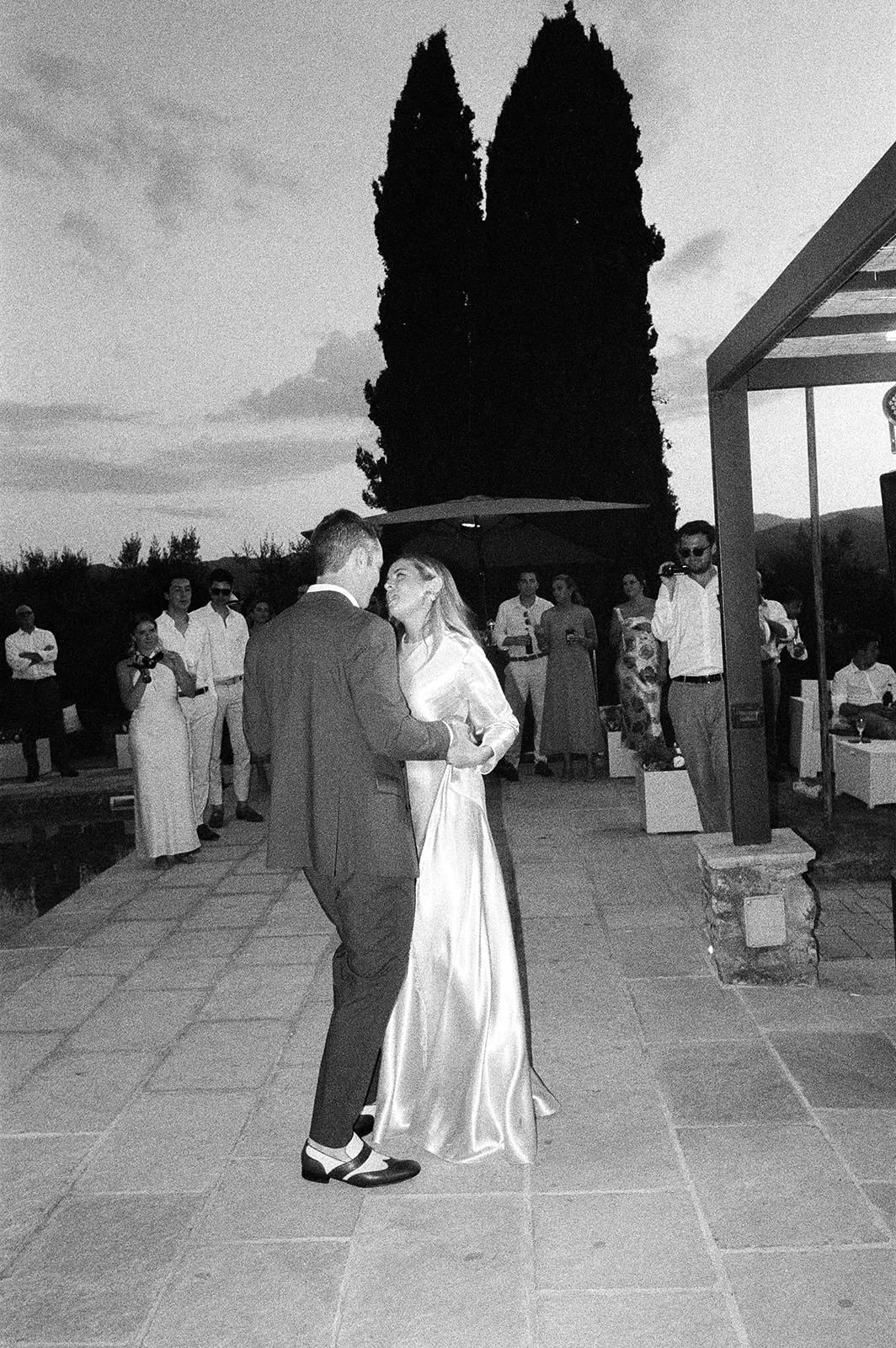 A bride in a long satin gown and veil dances with a groom in a suit on an outdoor patio at dusk. Guests in formal attire watch, some smiling. Two tall trees and a pergola frame the scene. The photo is black and white. Tuscan Wedding Photographer