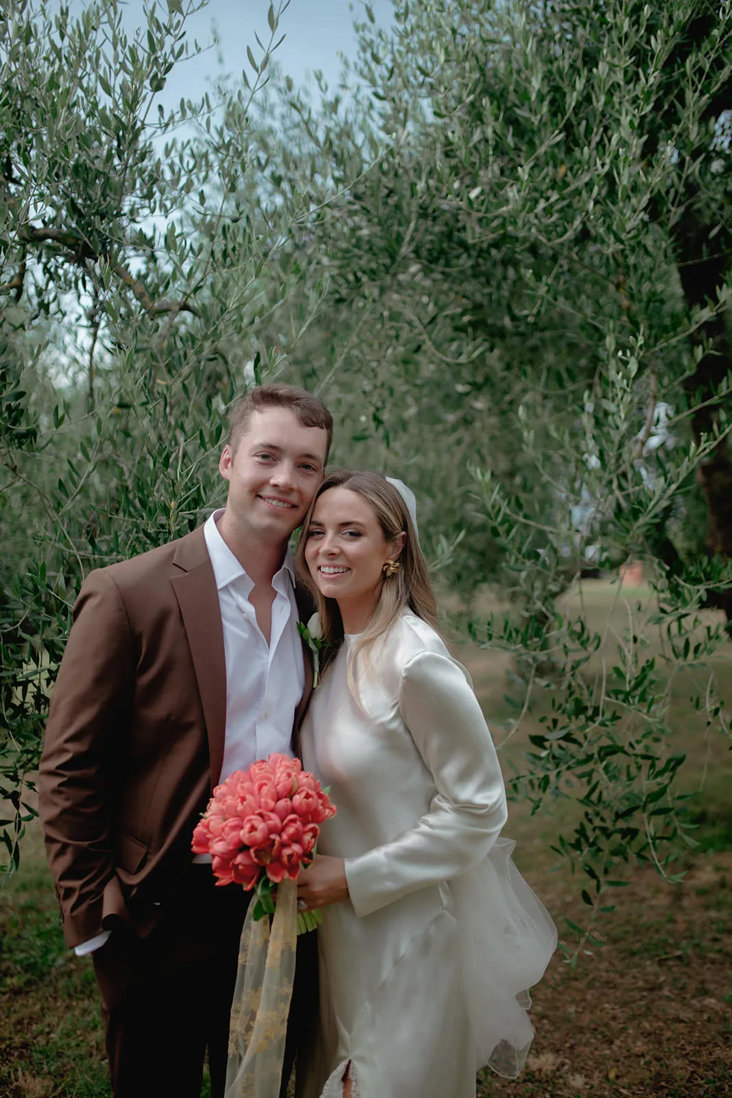 A smiling couple stands together outdoors, posing for a photo. The man wears a brown suit and white shirt, while the woman wears a white dress and holds a bouquet of pink flowers. Green leafy branches fill the soft-focus background. Tuscan Wedding Photographer