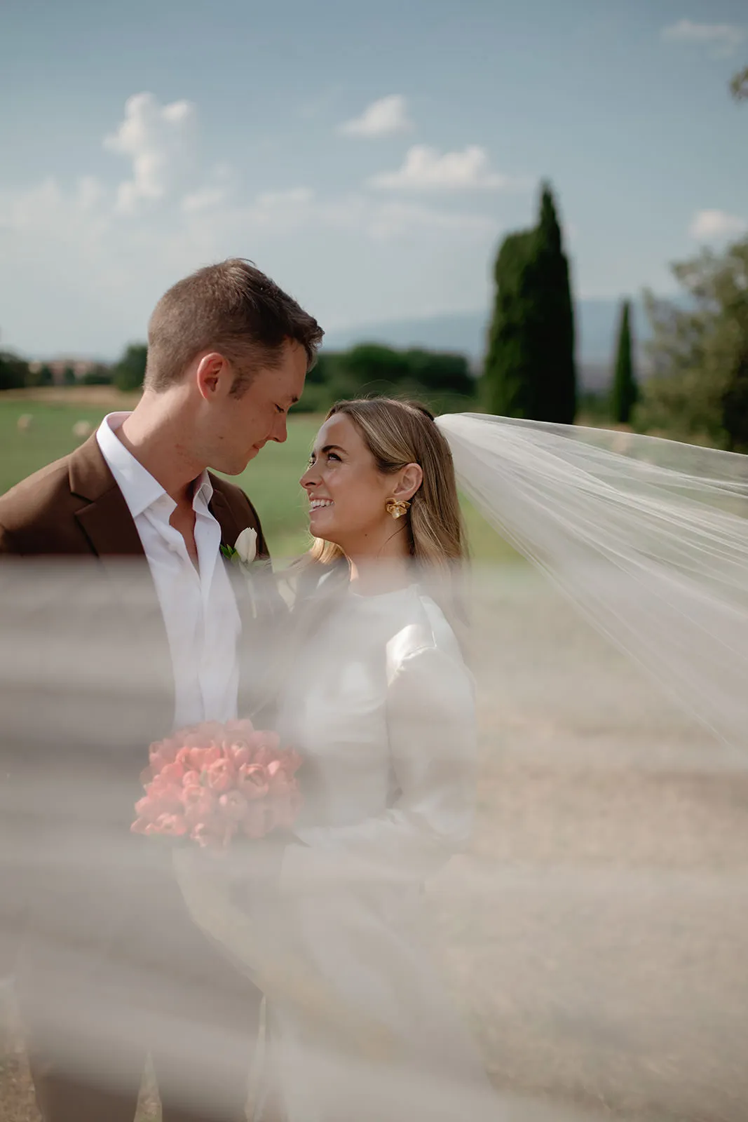 A bride and groom gaze into each other’s eyes outdoors. The bride holds pink flowers and wears a long flowing veil. The groom wears a brown jacket and white shirt. The scene is softly blurred in the foreground, with greenery and trees in the background. Tuscan Wedding Photographer