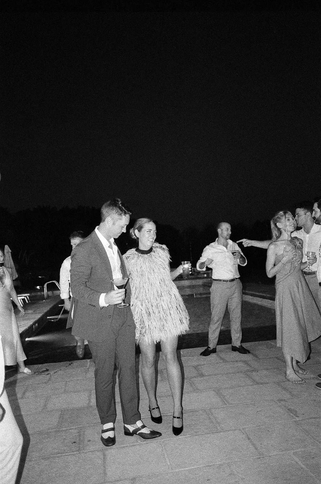 A group of people dressed in formal attire stand and mingle by a pool at night. In the foreground, a man in a suit and a woman in a fringed dress hold drinks and talk. The scene is lit softly, with others chatting in the background. Tuscan Wedding Photographer