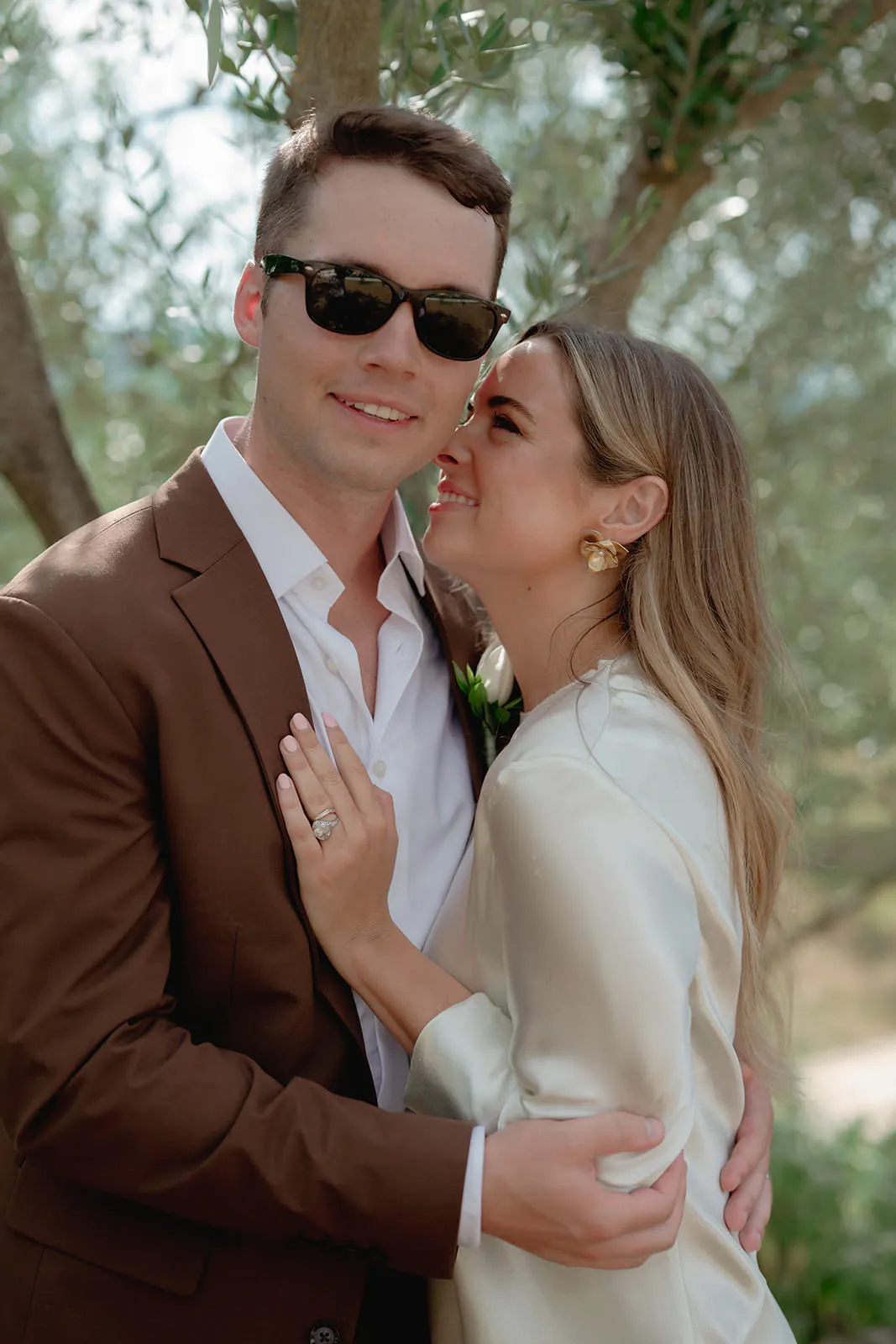 A man in a brown suit and sunglasses hugs a smiling woman in a white dress. She holds his chest and shows off a ring, leaning in close to his face, possibly whispering. They stand outdoors with trees and sunlight in the background. Tuscan Wedding Photographer