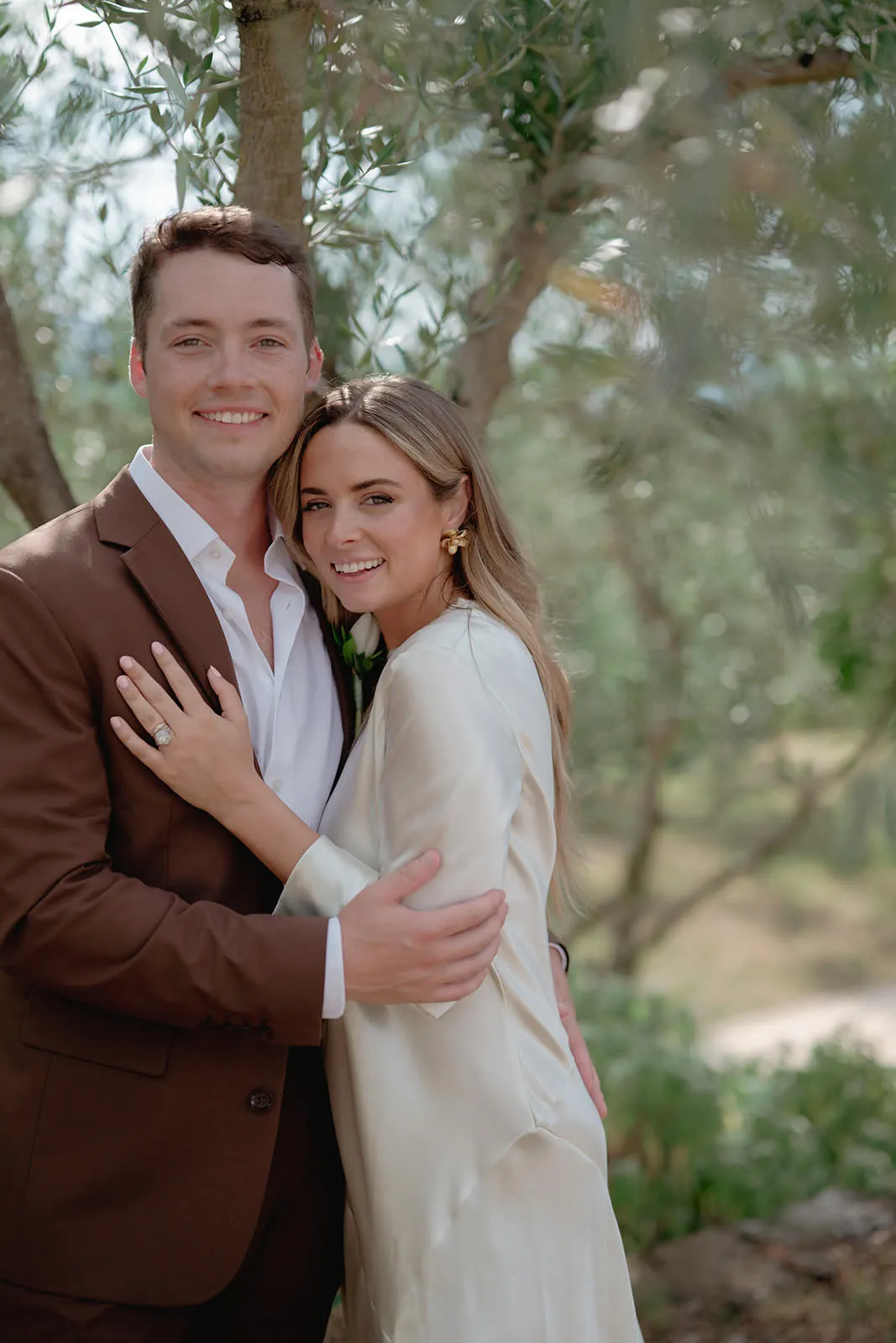 A smiling couple poses outdoors, standing close with arms around each other. The man wears a brown suit and white shirt, while the woman wears a light cream dress. Trees and greenery form a soft, natural background. Both look happy and relaxed. Tuscan Wedding Photographer