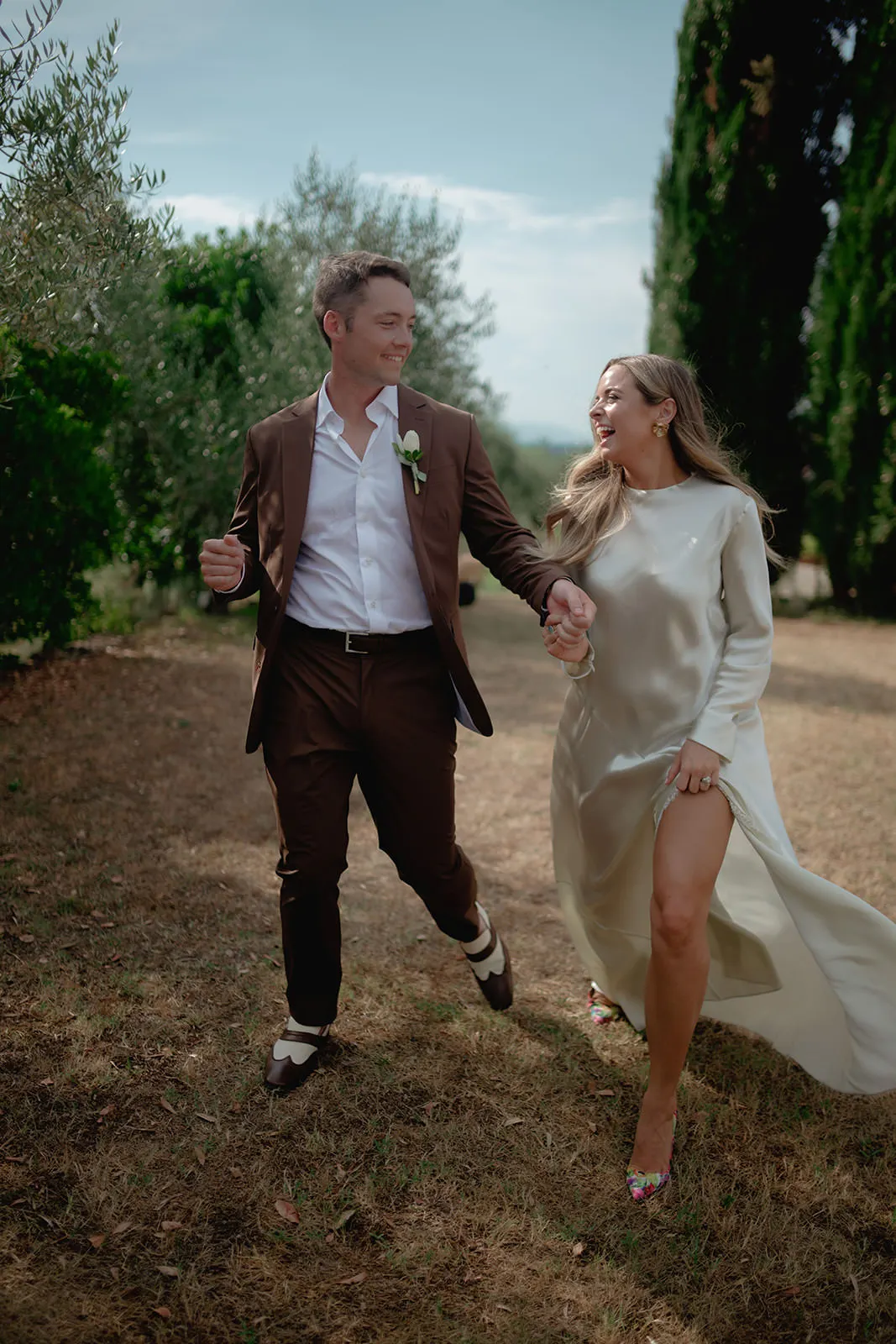A smiling couple walks hand-in-hand outdoors. The man wears a brown suit with a white shirt and boutonnière, and the woman wears a long, light-colored dress with a high slit. They appear joyful, surrounded by green trees and grass. Tuscan Wedding Photographer
