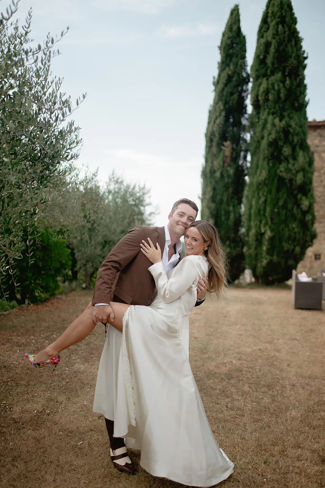 A smiling man in a brown suit playfully lifts a smiling woman in a long white dress outdoors. They pose on dry grass with tall green trees and a stone building in the background, creating a joyful, celebratory atmosphere. Tuscan Wedding Photographer