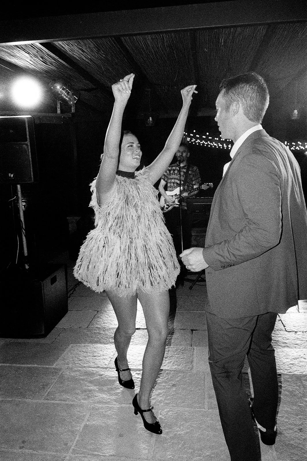 A woman in a textured, fringed dress dances with arms raised, smiling, while facing a man in a suit. String lights and a musician playing guitar are visible in the background, creating a lively, festive atmosphere. Tuscan Wedding Photographer
