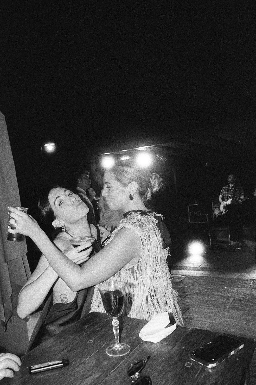 Two women pose and laugh at a table with drinks in hand at a dimly lit party. One puckers her lips toward the other. There are wine glasses, a phone, and napkins on the table. In the background, a musician performs on stage under bright lights. Tuscan Wedding Photographer