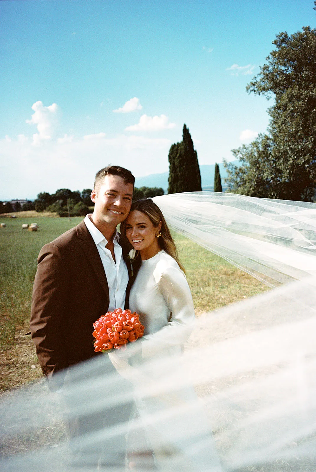 A newlywed couple smiles outdoors on a sunny day. The groom wears a brown suit; the bride, in white, holds an orange bouquet. Her veil flows in the wind. Greenery, trees, and a blue sky are in the background. Tuscan Wedding Photographer