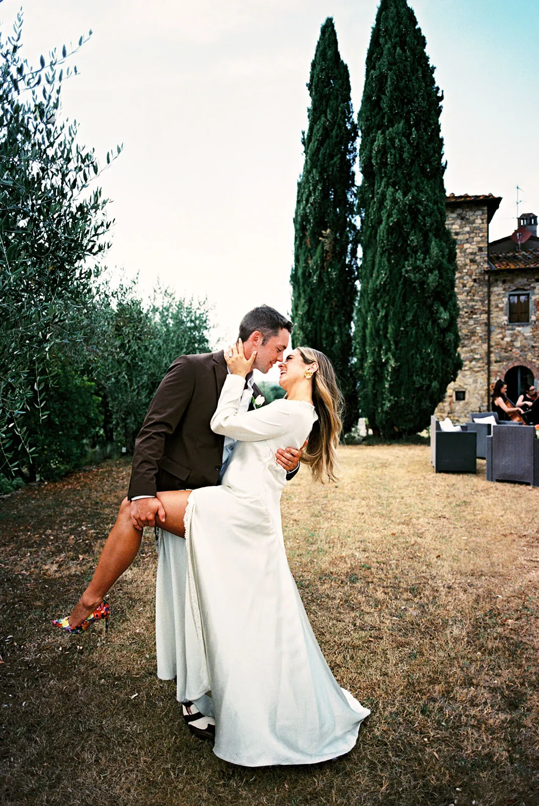 A couple kisses outdoors; the groom in a brown suit lifts the bride in a long white dress. Tall green trees and a stone house are in the background. The lawn is dry with some wicker furniture visible on the right. Tuscan Wedding Photographer