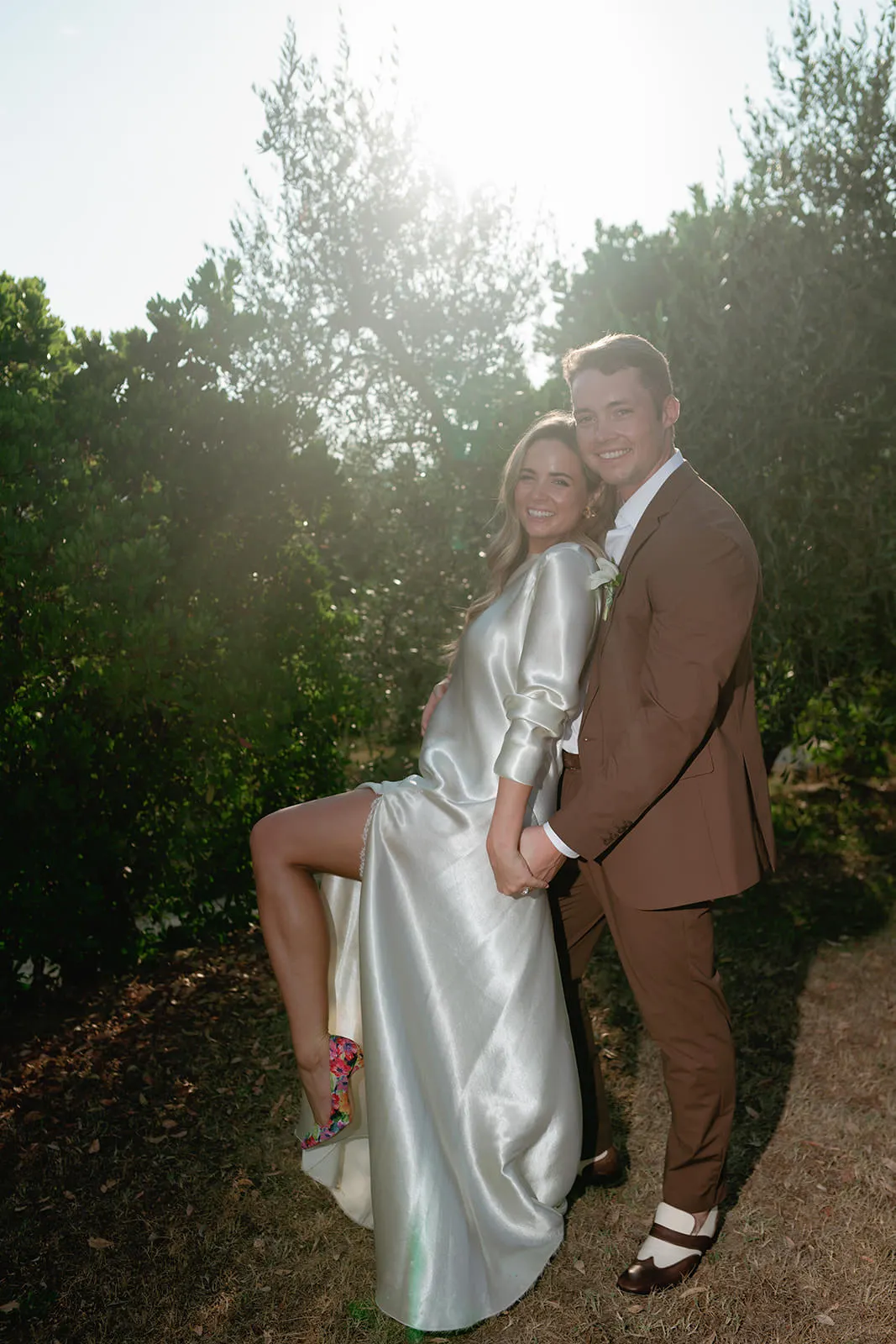 A smiling couple poses outdoors in sunlight. The woman wears a shiny silver dress with a high slit and colorful heels, lifting one leg playfully. The man stands behind her in a brown suit and white shirt. Greenery surrounds them, and both look happy and relaxed. Tuscan Wedding Photographer