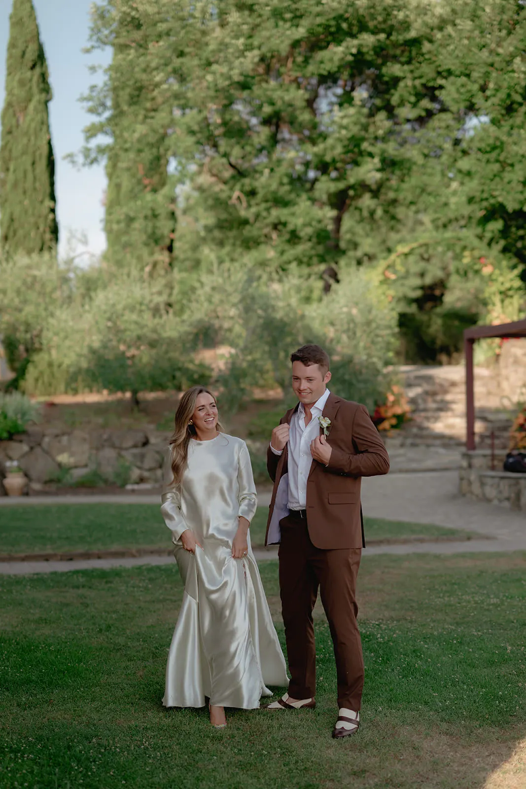 A woman in a long, silky, off-white dress stands beside a man in a brown suit with a white shirt and boutonnière. They smile at each other outdoors, surrounded by grass, stone walls, and lush green trees under soft daylight. Tuscan Wedding Photographer