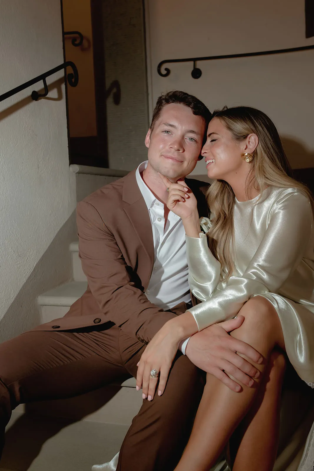 A couple sits on indoor stairs. The man wears a brown suit and white shirt, smiling at the camera. The woman, in a shiny cream dress, leans affectionately on him, smiling with eyes closed and touching his chin, her engagement ring visible. Warm, soft lighting. Tuscan Wedding Photographer