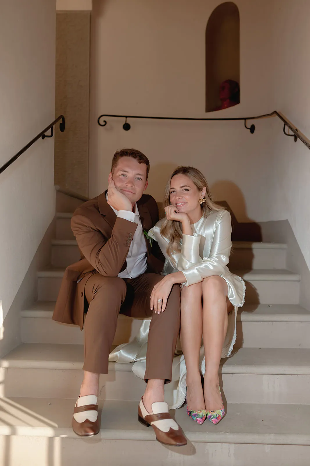 A smiling man in a brown suit and a smiling woman in a pale satin dress sit close together on indoor stairs, resting their chins on their hands. Both are barefoot; her colorful heels and his brown loafers sit on the step below. Sunlight casts soft shadows. Tuscan Wedding Photographer