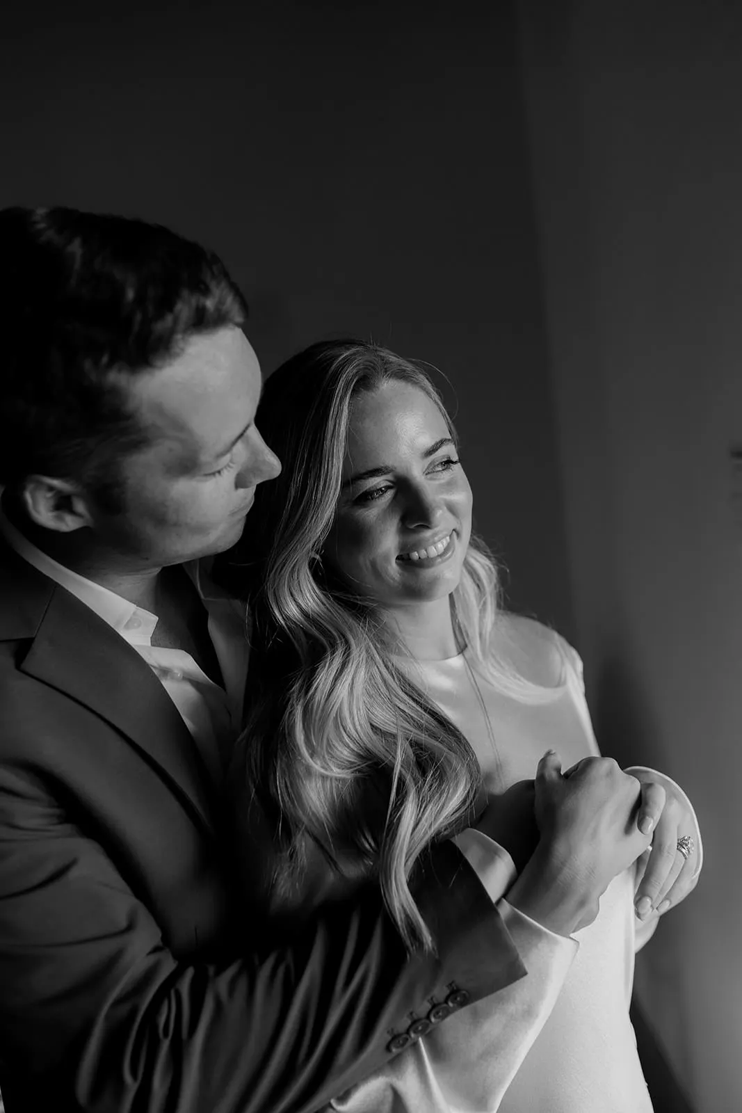 A man in a suit lovingly embraces a smiling woman from behind. The woman has long, wavy hair and holds the man’s arms. Both look content, standing in soft, natural light against a plain background. The photo is black and white. Tuscan Wedding Photographer