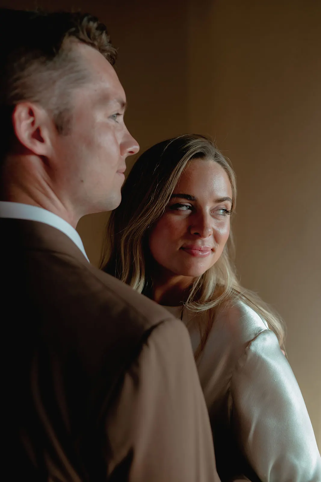 A man in a brown suit stands beside a woman in a silky white blouse. The woman, with long blonde hair, gazes at the man with a gentle, affectionate expression. The background is softly lit, creating a warm, intimate atmosphere. Tuscan Wedding Photographer