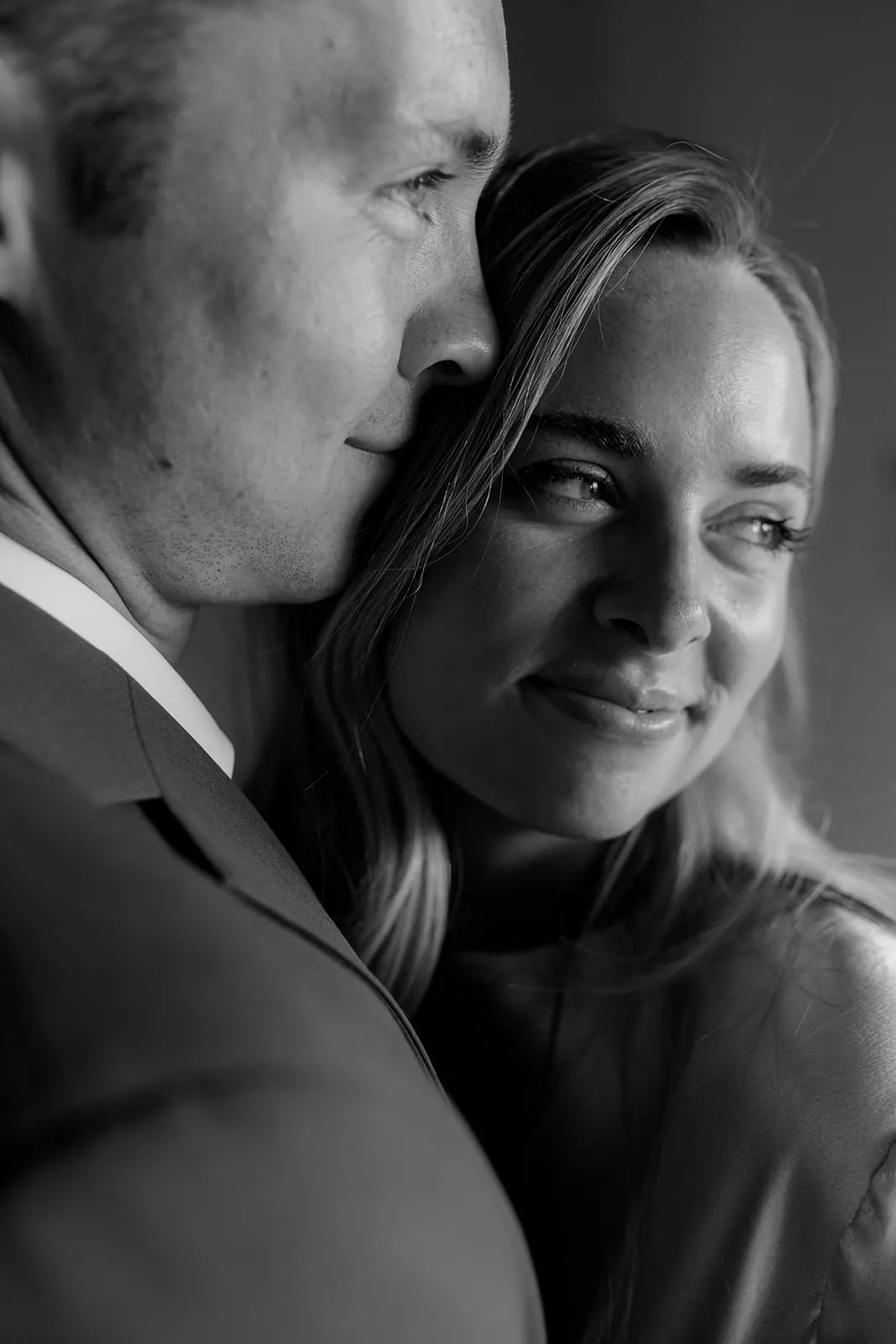 A close-up black-and-white photo of a couple. The man, in a suit, gently leans his face near the womans forehead. The woman smiles subtly, looking slightly away. Both appear content and relaxed, captured in soft, natural light. Tuscan Wedding Photographer