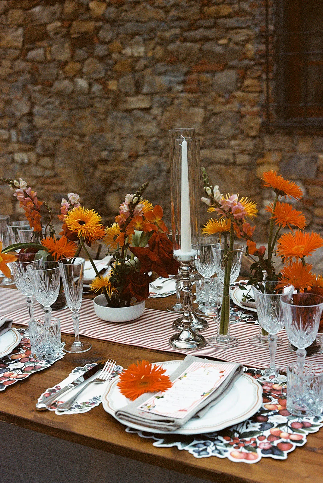 Elegant outdoor table set against a stone wall, decorated with orange and yellow flowers, glassware, silver cutlery, floral placemats, folded napkins, and a tall white candle in a silver holder at the center. Festive and inviting atmosphere. Tuscan Wedding Photographer