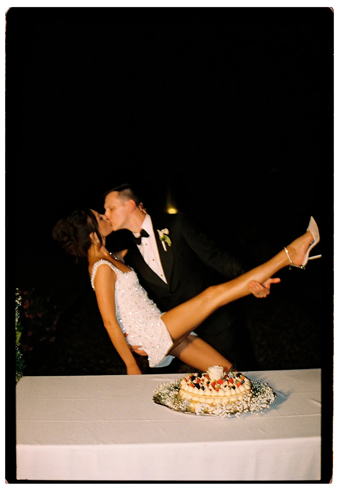A couple dressed in formal attire passionately kiss in front of a table with a decorated cake. The woman, wearing a short white dress and heels, lifts one leg playfully as the man, in a black suit and bow tie, supports her. The background is dark. Tuscan Wedding Photographer