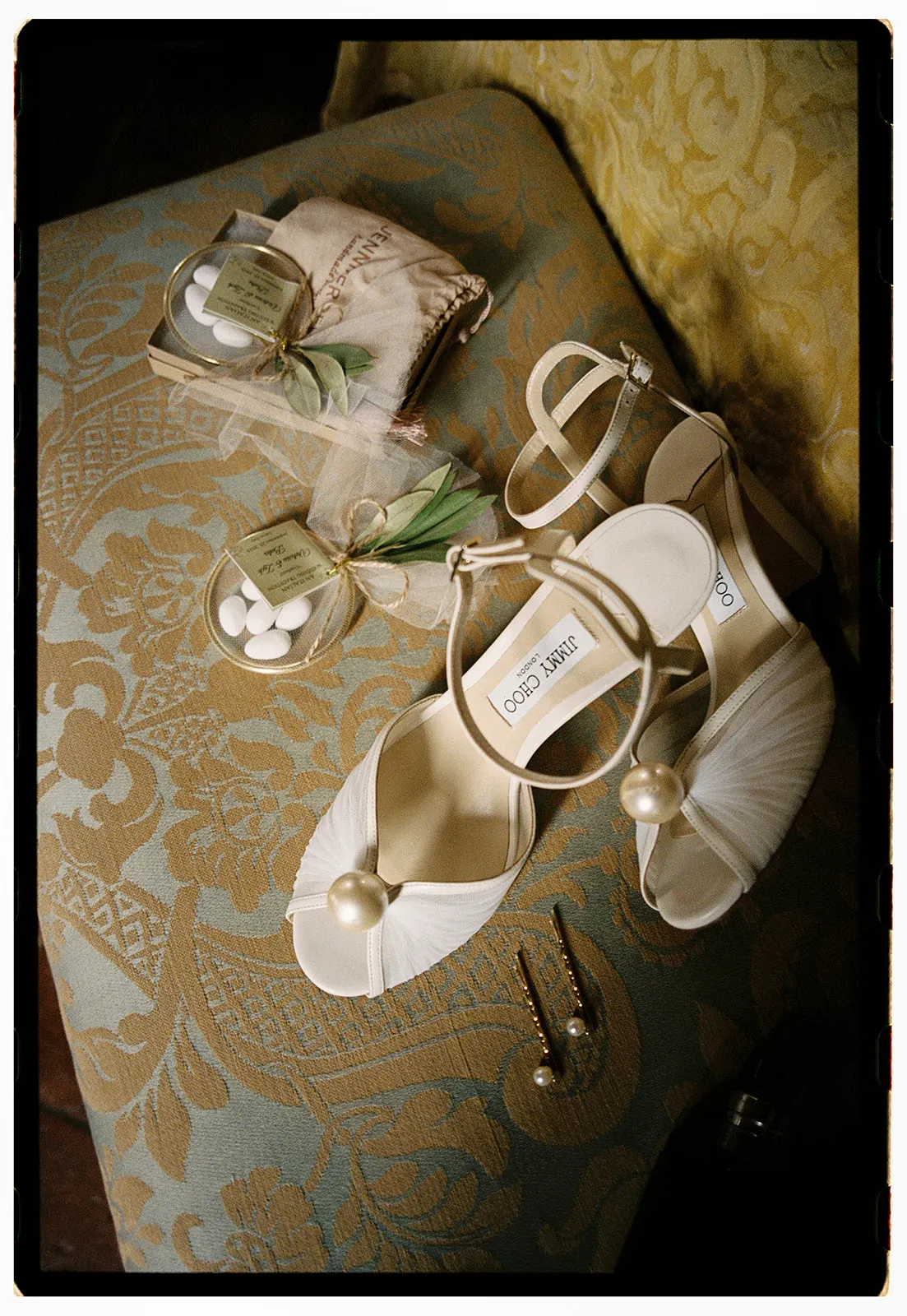 A pair of elegant white Jimmy Choo heels with pearl accents sits on a light blue and gold patterned bench, alongside a small beige pouch, boxes of sugared almonds tied with green ribbon, and two gold hairpins. Tuscan Wedding Photographer