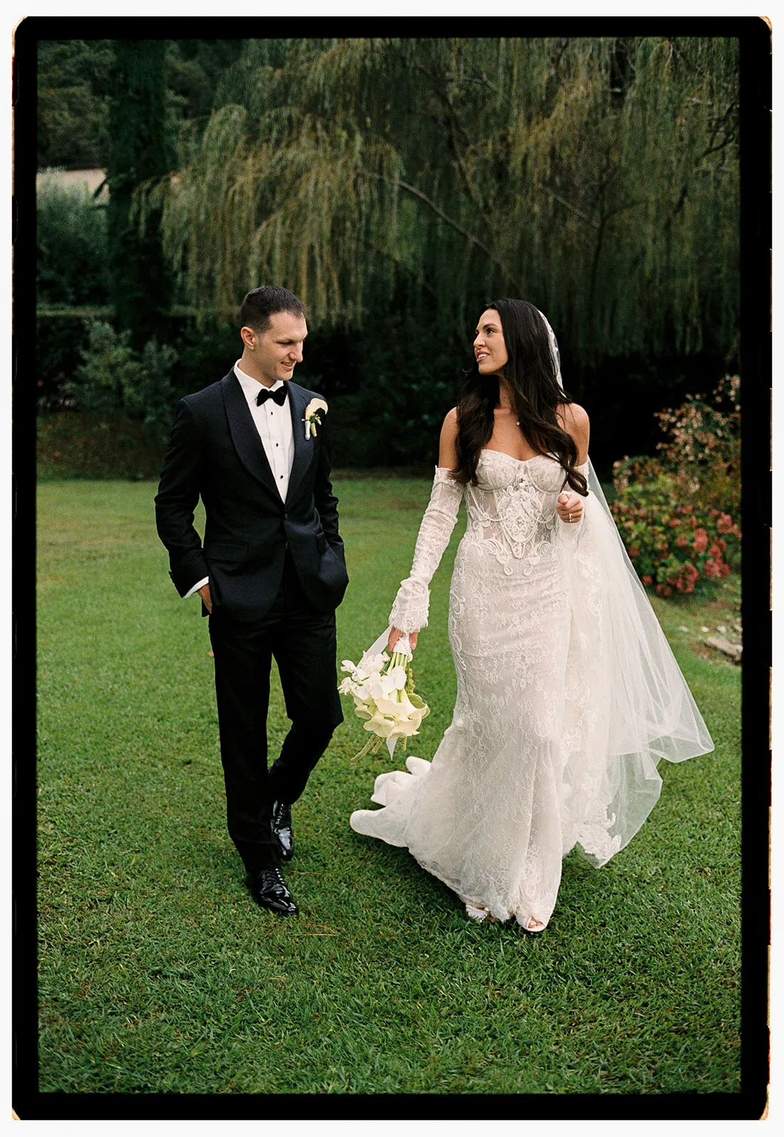 A bride in a lacy, off-shoulder white gown holds a bouquet and walks beside a groom in a black tuxedo on a grassy lawn. Both smile at each other, surrounded by greenery and flowers, with trees and bushes in the background. Tuscan Wedding Photographer