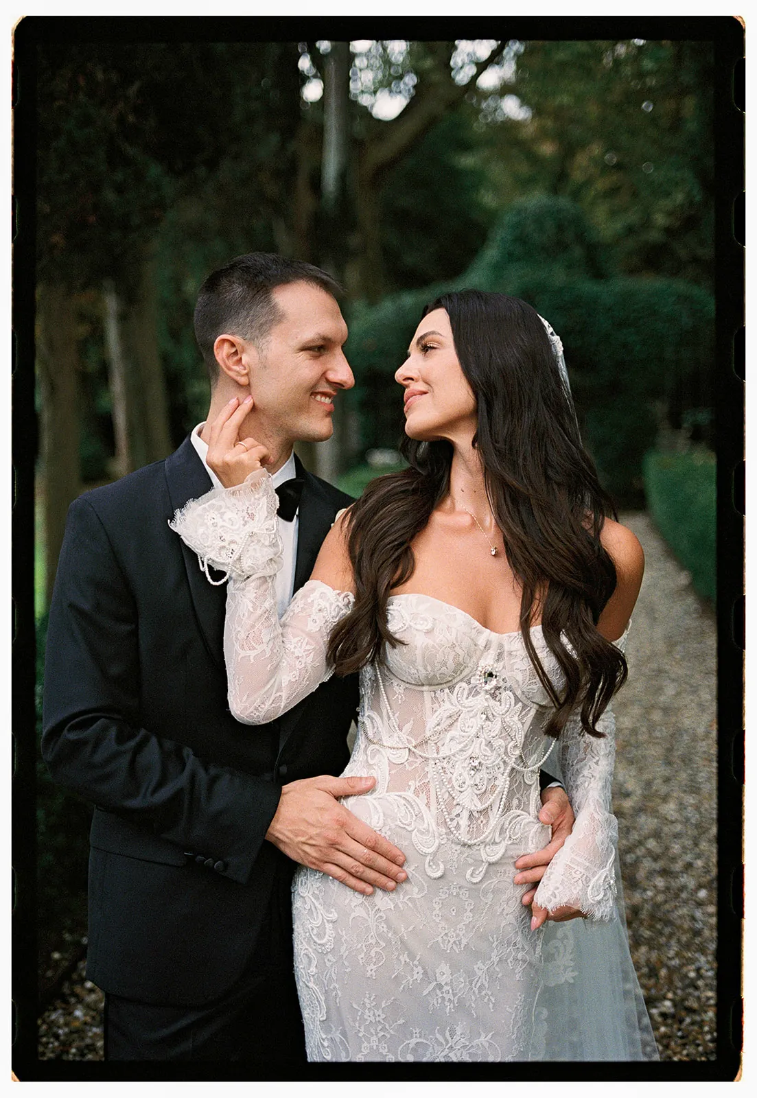 A bride and groom stand close together outdoors, smiling at each other. The bride wears a white, lace, off-the-shoulder wedding gown with long sleeves, while the groom wears a classic black tuxedo. Lush greenery and a stone path are visible in the background. Tuscan Wedding Photographer