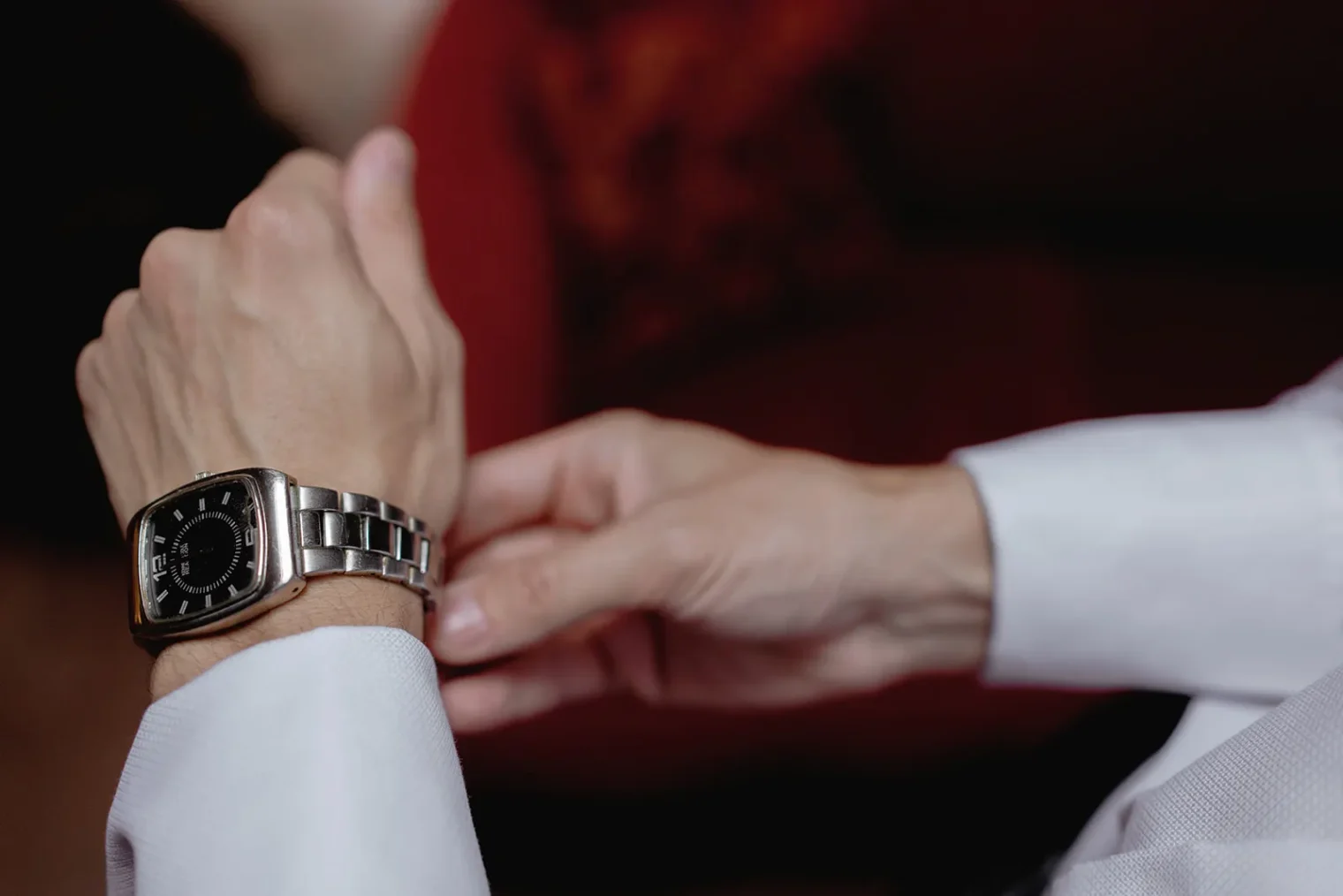 A close-up of a person’s hands adjusting a silver wristwatch with a black face. The person is wearing a white long-sleeve shirt, and the background is blurred with shades of red and brown. Tuscan Wedding Photographer